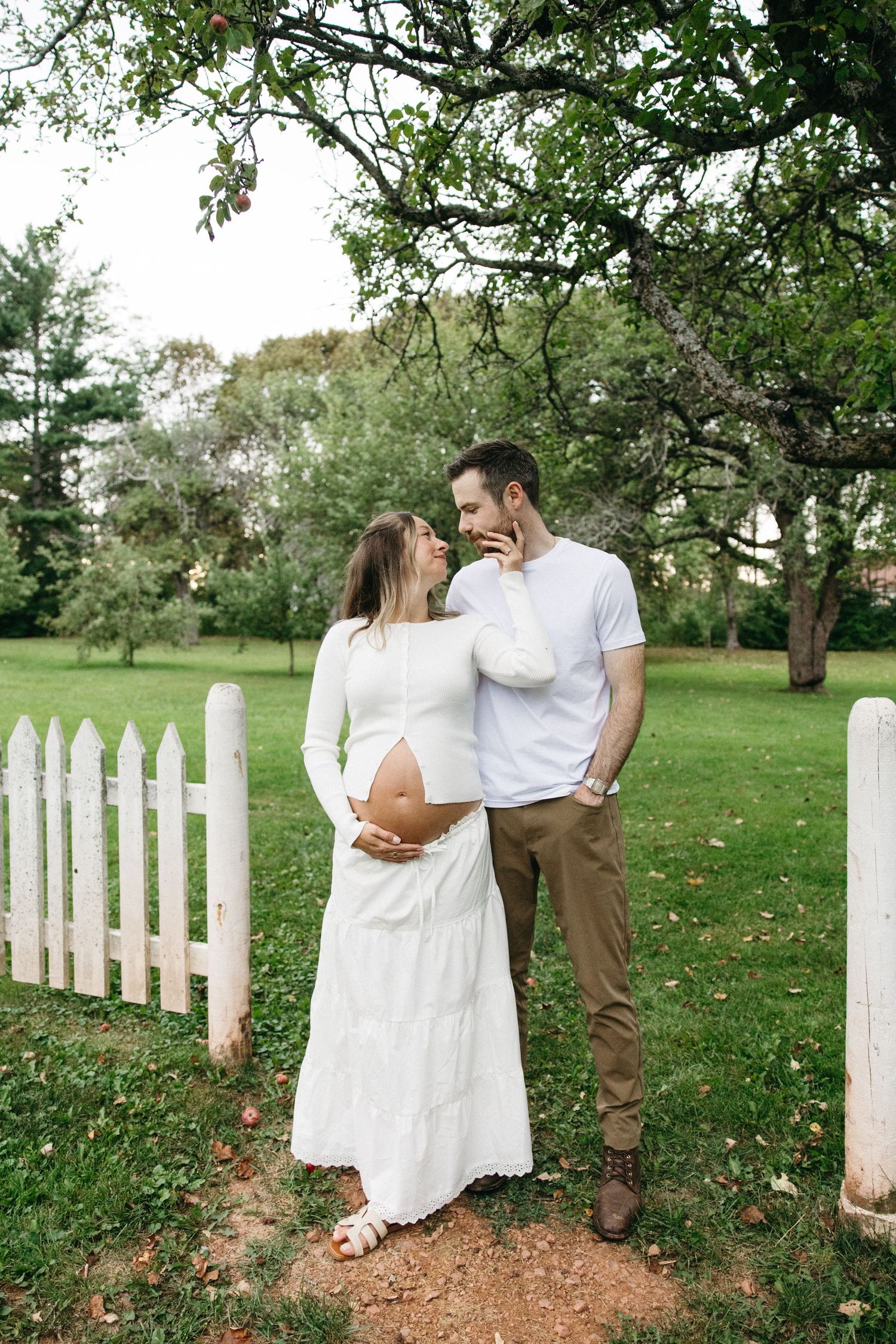 Expecting couple in white outfits share tender moment by picket fence in green park setting.