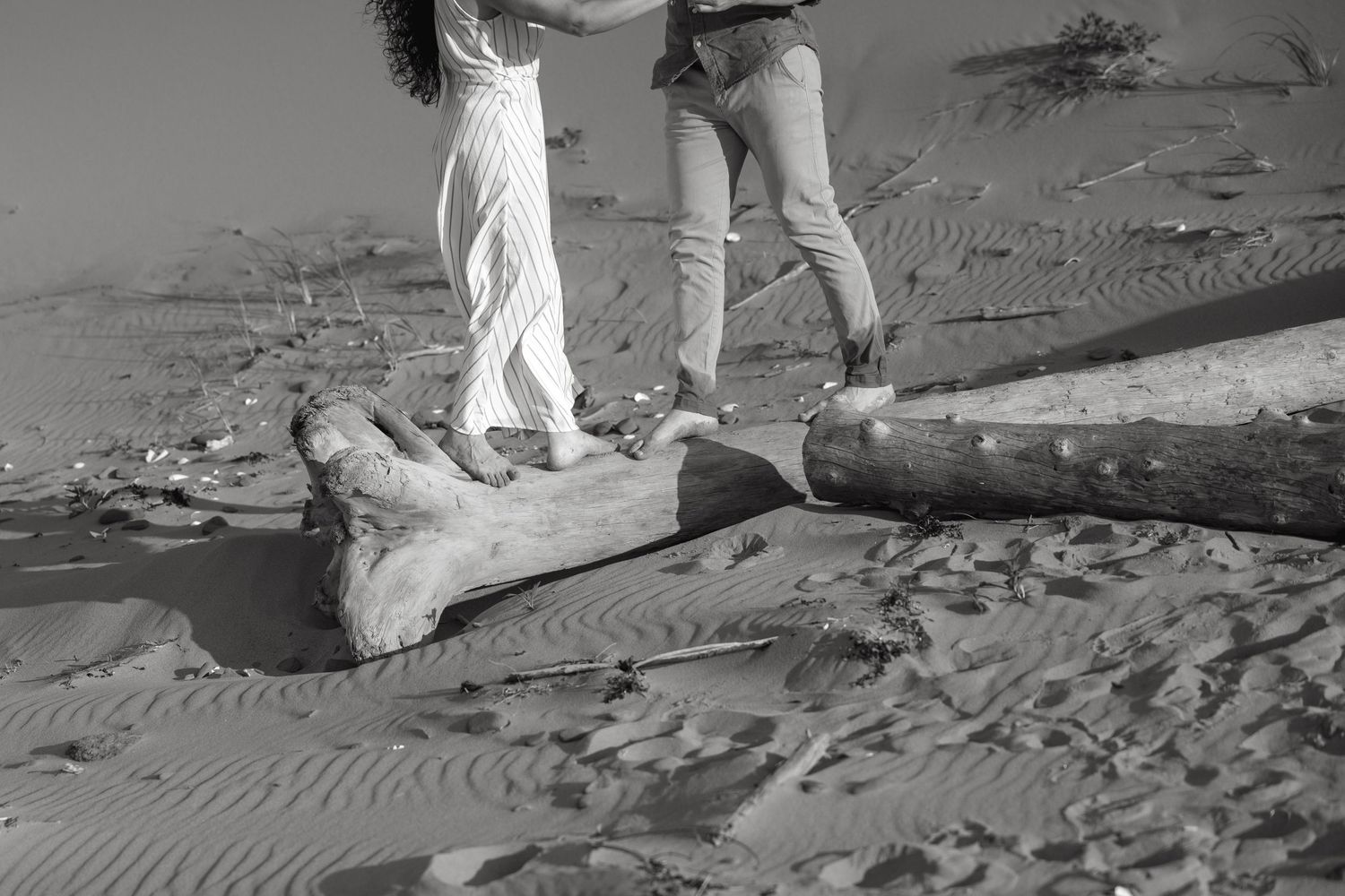 Black and white photo of a couple standing on driftwood logs in sandy desert landscape.