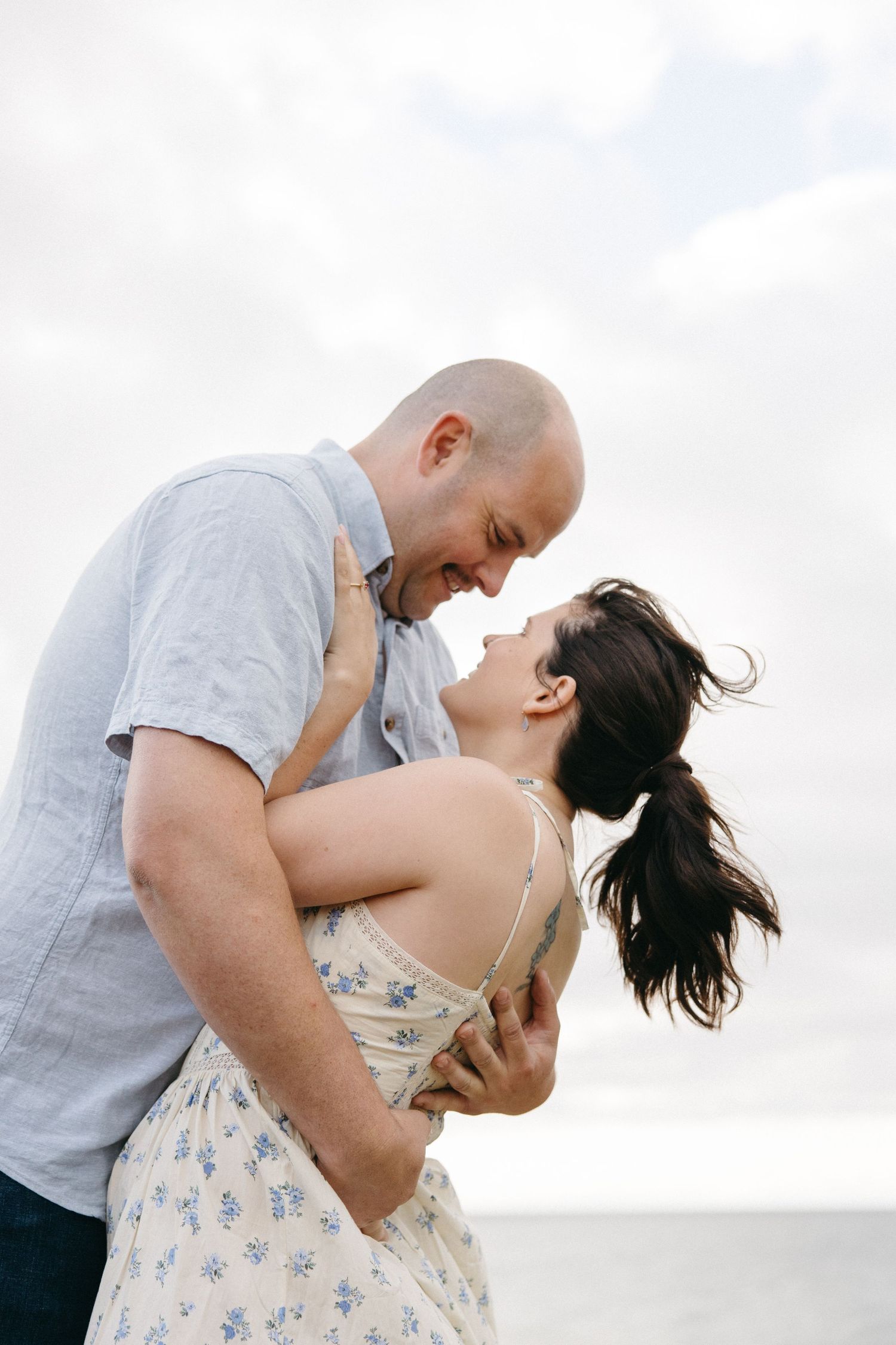 A laughing couple embraces and nearly kisses against a bright cloudy sky at the beach.