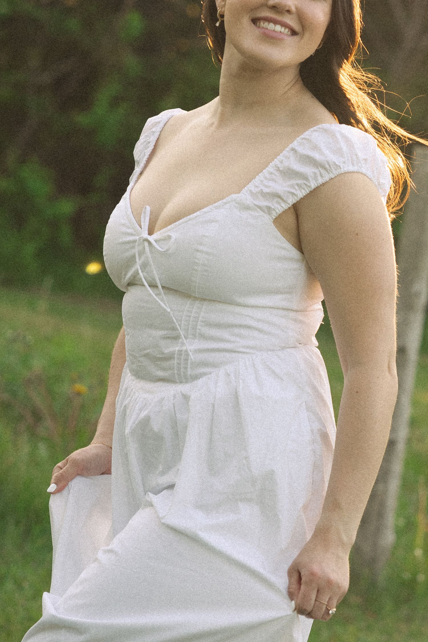 Woman in white sweetheart neckline dress smiling outdoors in a lush green garden setting.