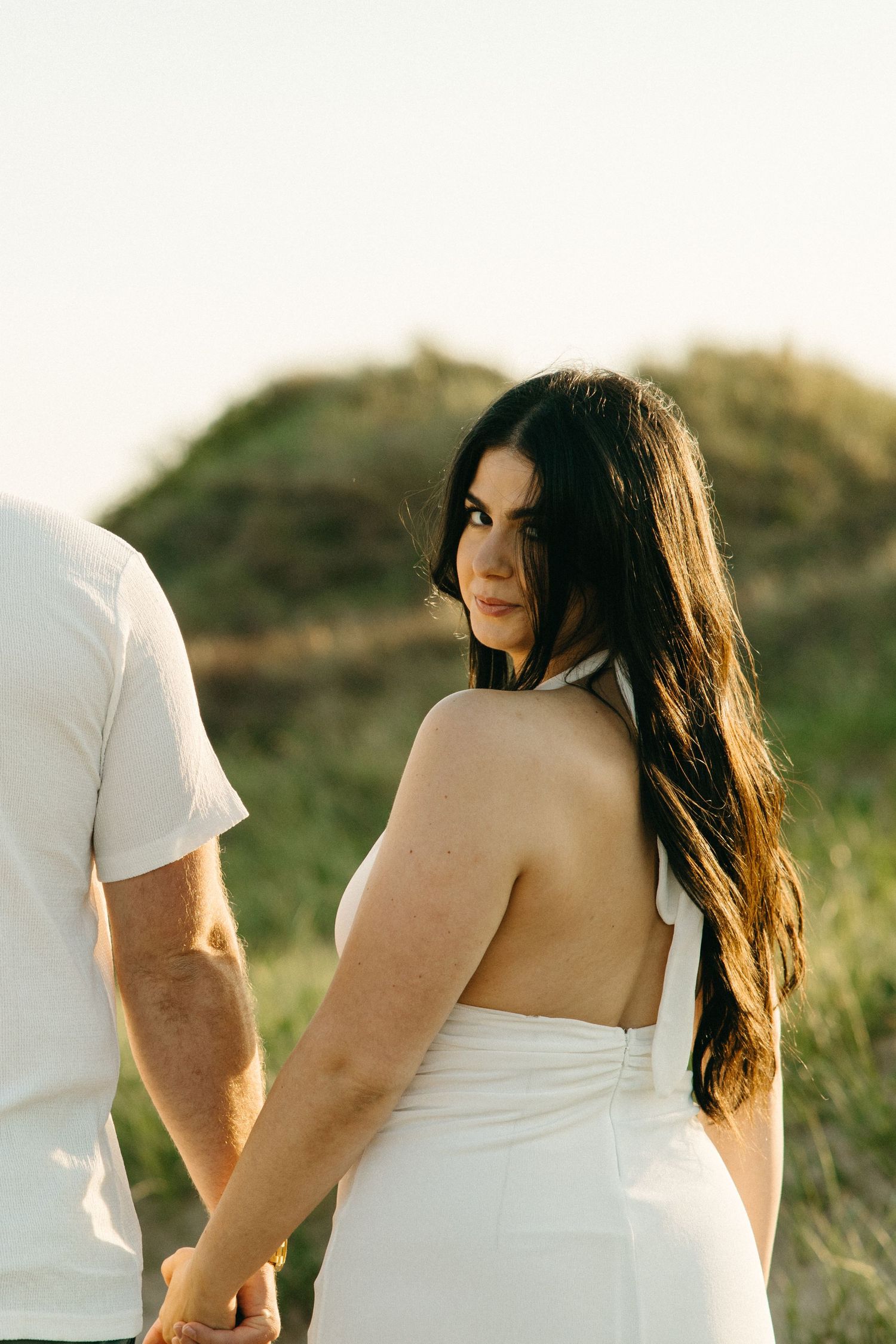 Woman in backless white halter dress looks over shoulder while holding hands with partner outdoors at golden hour.