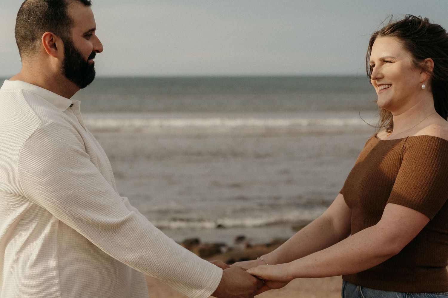 A couple holds hands and smiles at each other on a beach with waves in the background.
