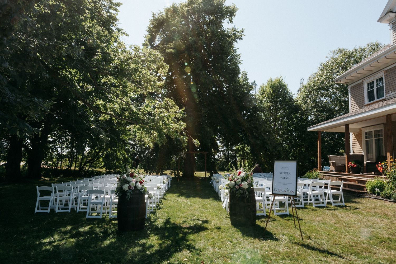 Outdoor wedding ceremony setup with white chairs on a sunny lawn in front of a house.
