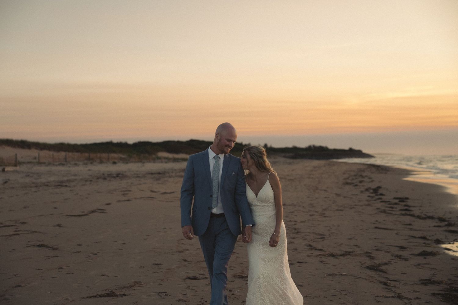 A couple walks along a scenic beach at sunset with golden light illuminating the sandy shoreline.