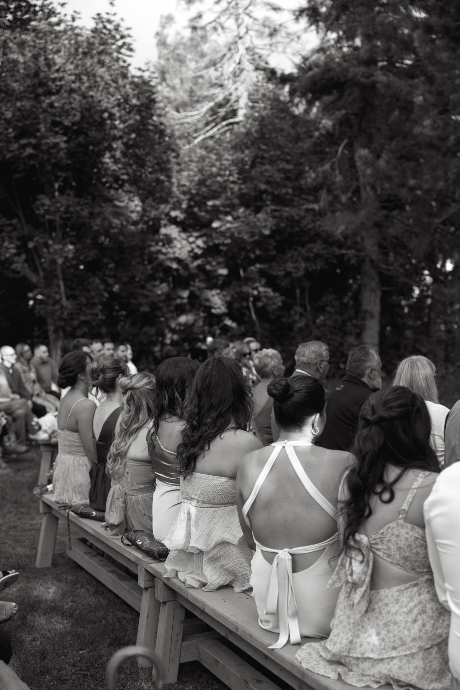Wedding guests seated on wooden benches during an outdoor ceremony in a wooded setting.