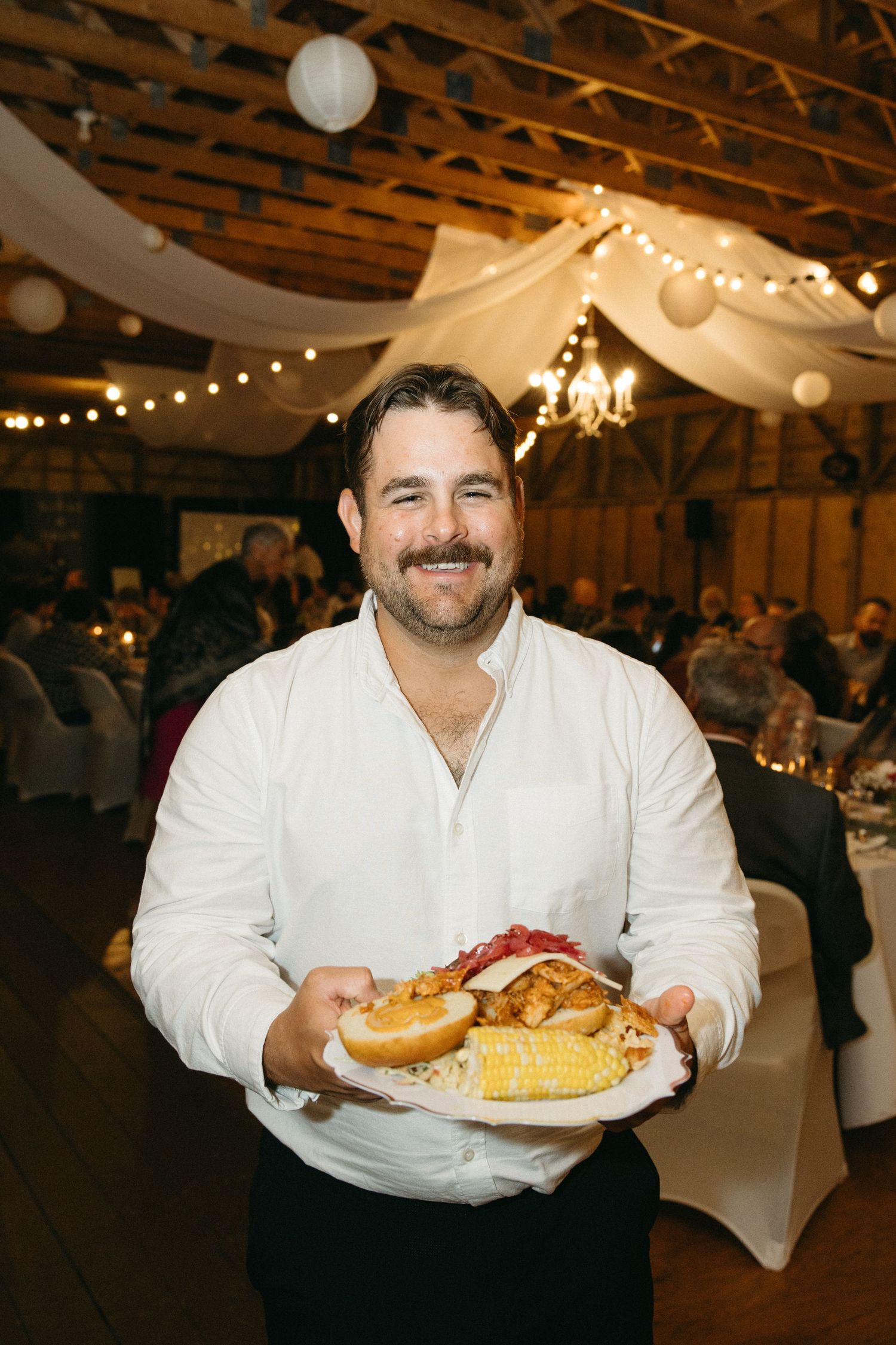 Guest in white shirt displays plates of food at a rustic wedding reception.