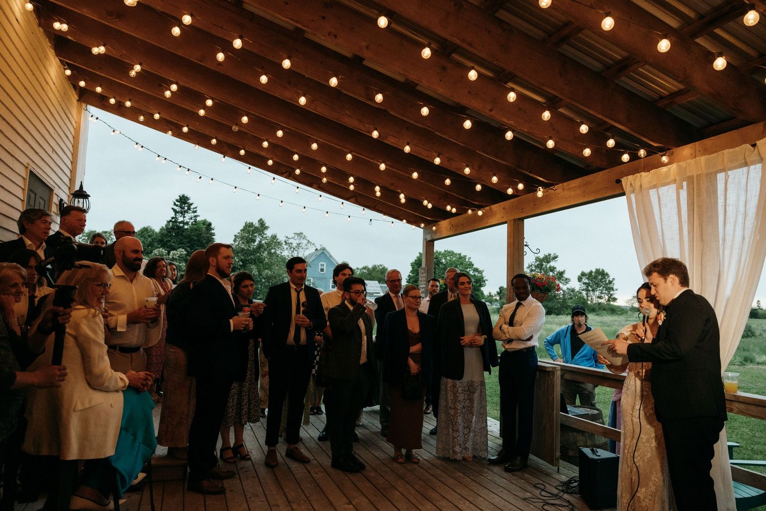 Wedding guests gather on a wooden deck under string lights during an outdoor reception.