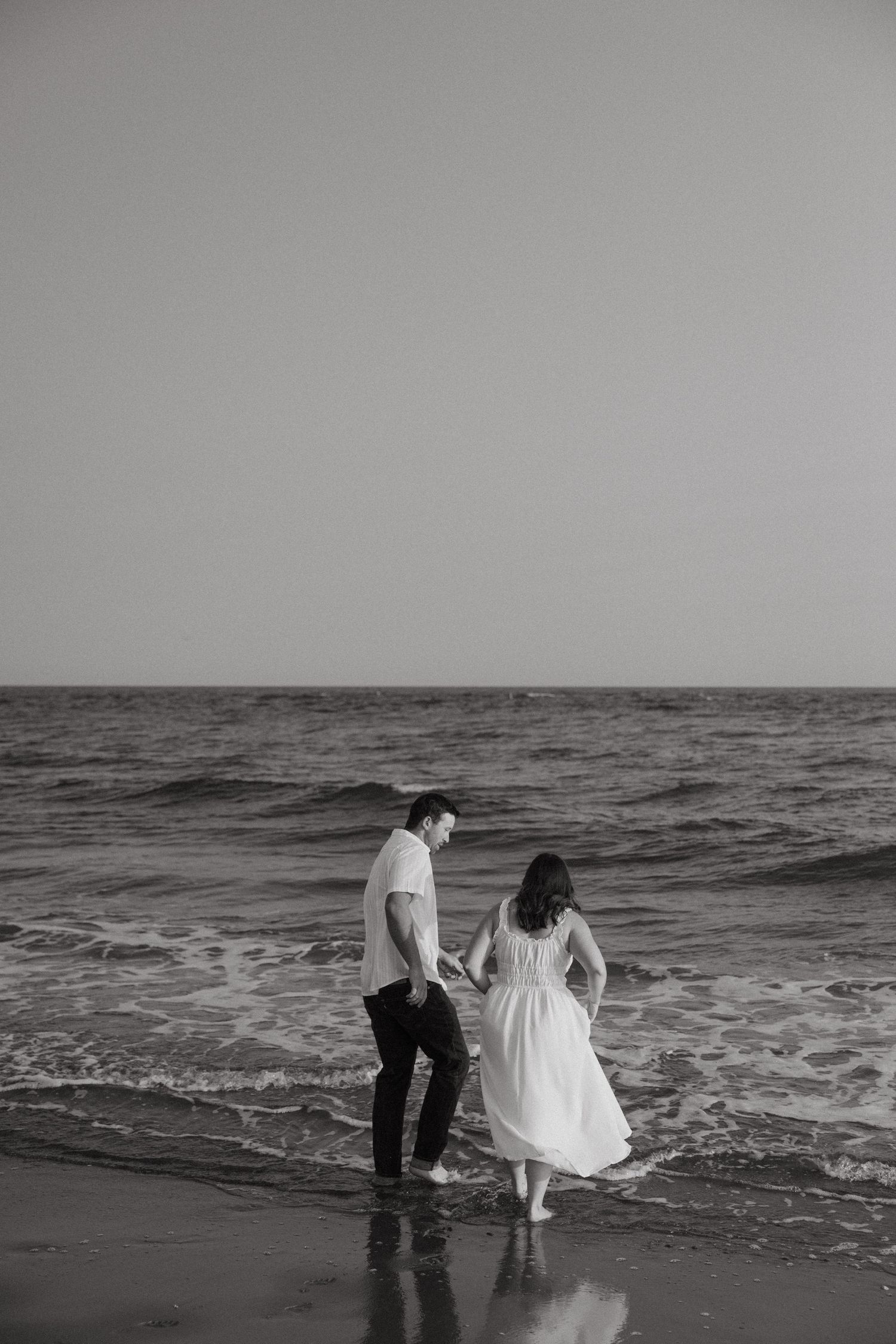A couple walks together in the ocean waves at the beach in black and white.