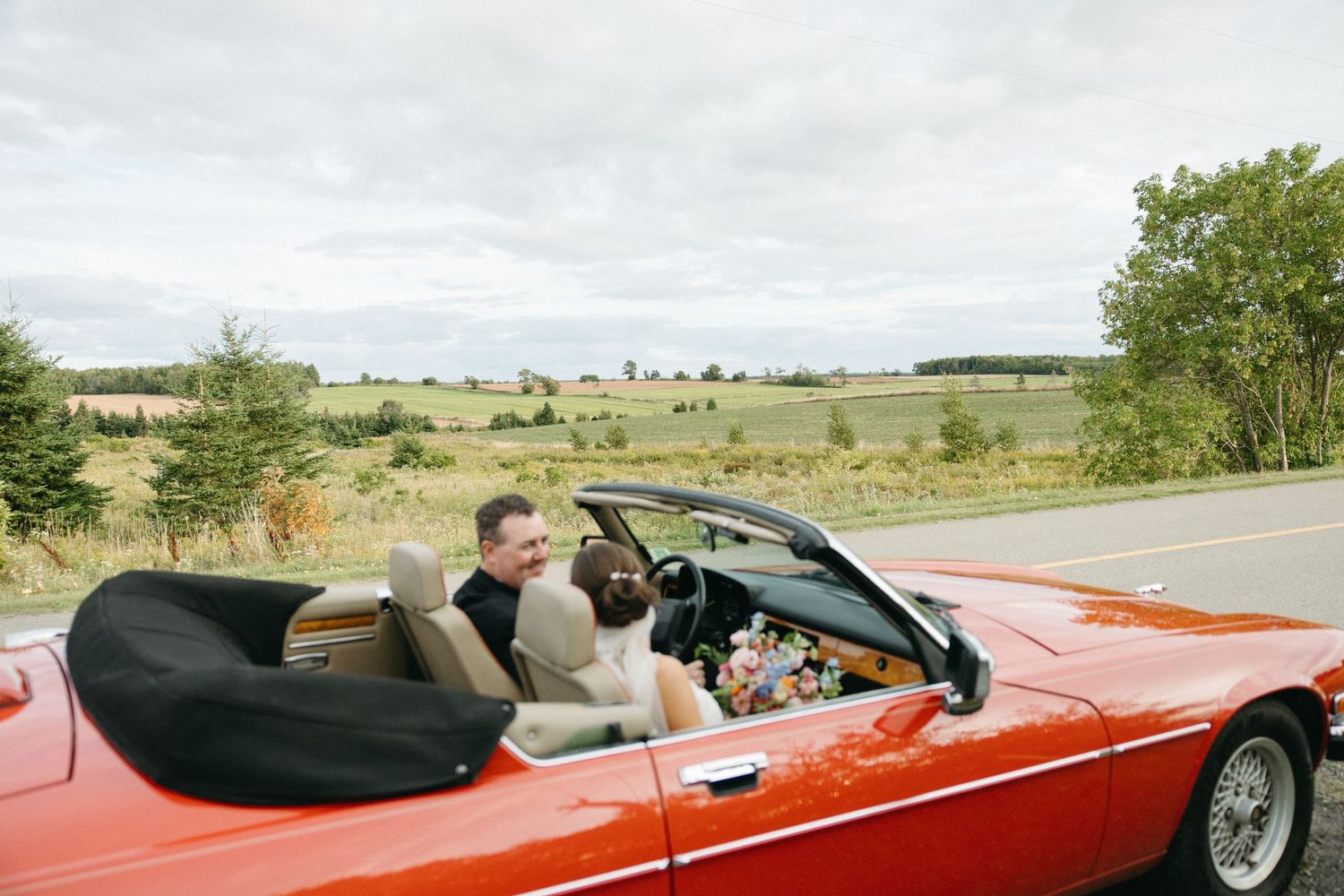 Red convertible car carrying wedding couple along rural road with open fields.