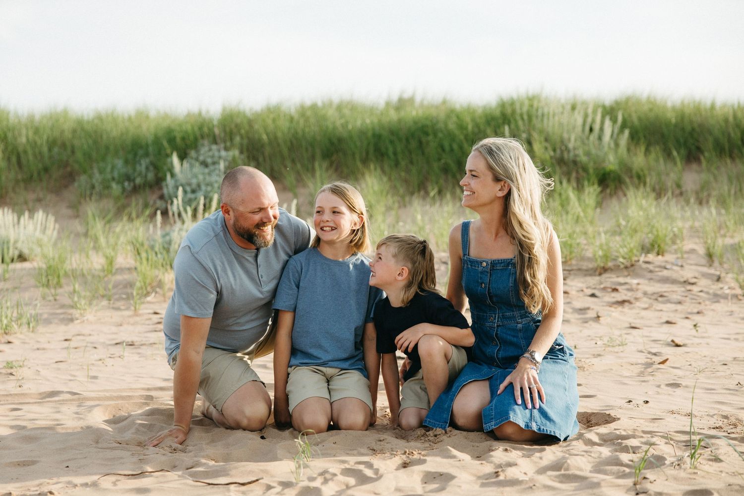 Smiling family of four sitting together on sandy beach with grass dunes behind.