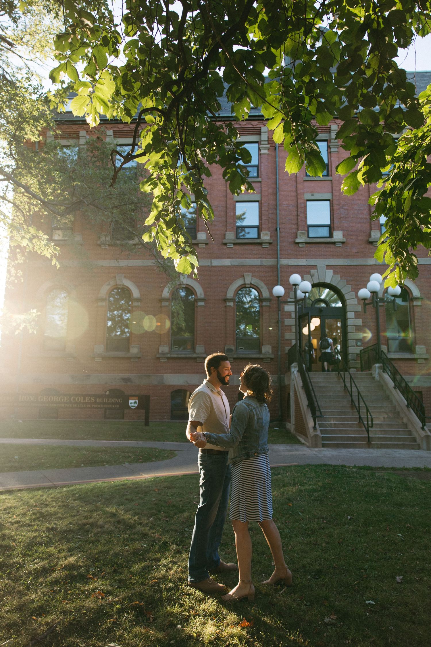 Two people dance and embrace on a lawn in front of a brick building during golden hour.