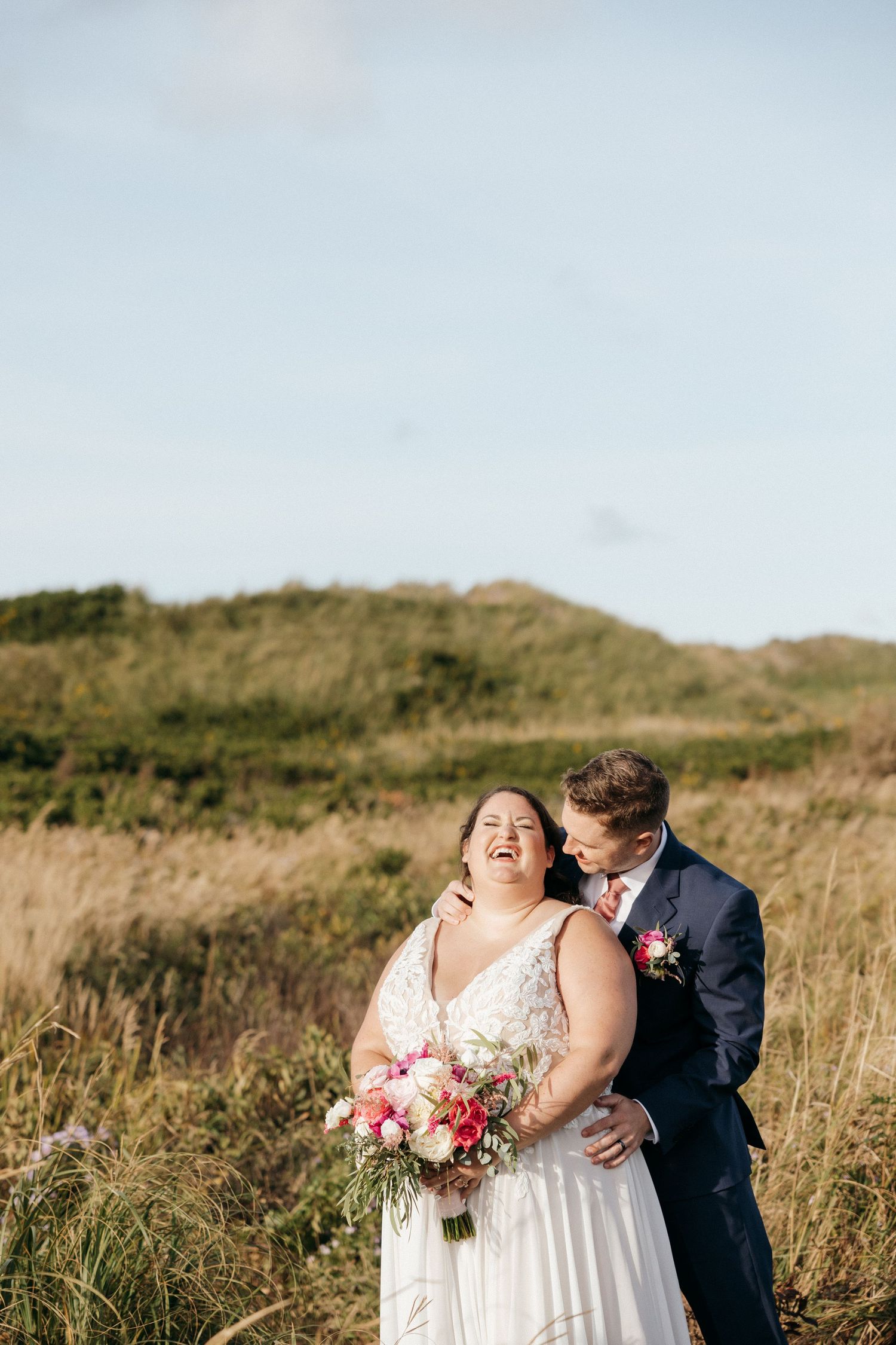 A couple embraces in a field during their wedding photoshoot with a bouquet of pink flowers.