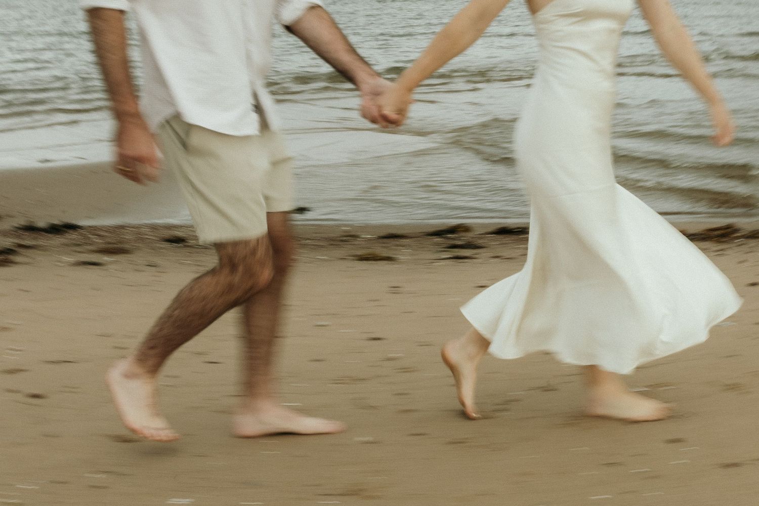 Cropped view of two people walking hand in hand on a beach, showing just their feet and flowing clothing.