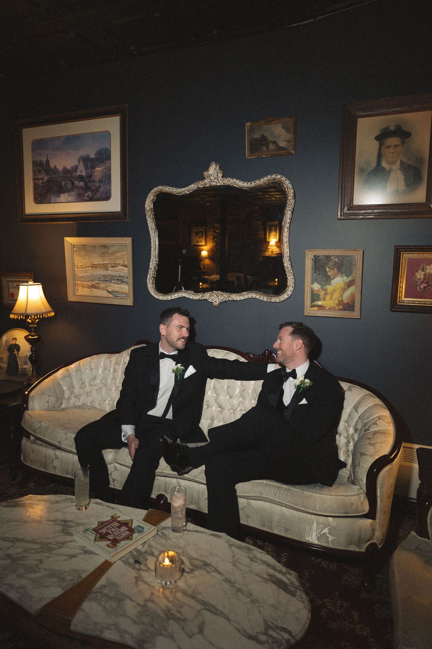 Two people in tuxedos lounging on an ornate white couch in a dark room with vintage wall art.