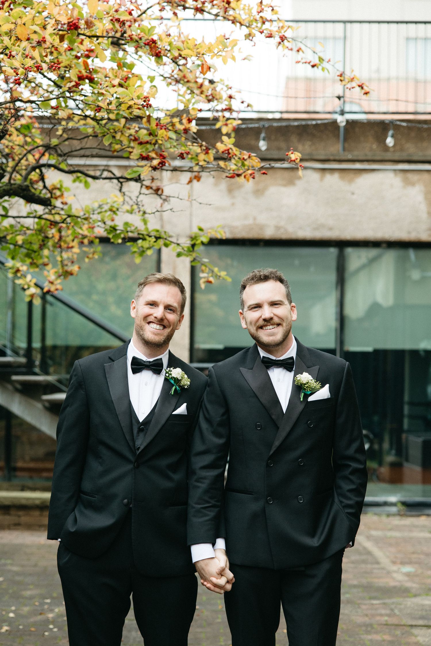 Two people in matching black tuxedos and bow ties hold hands while standing under autumn foliage.