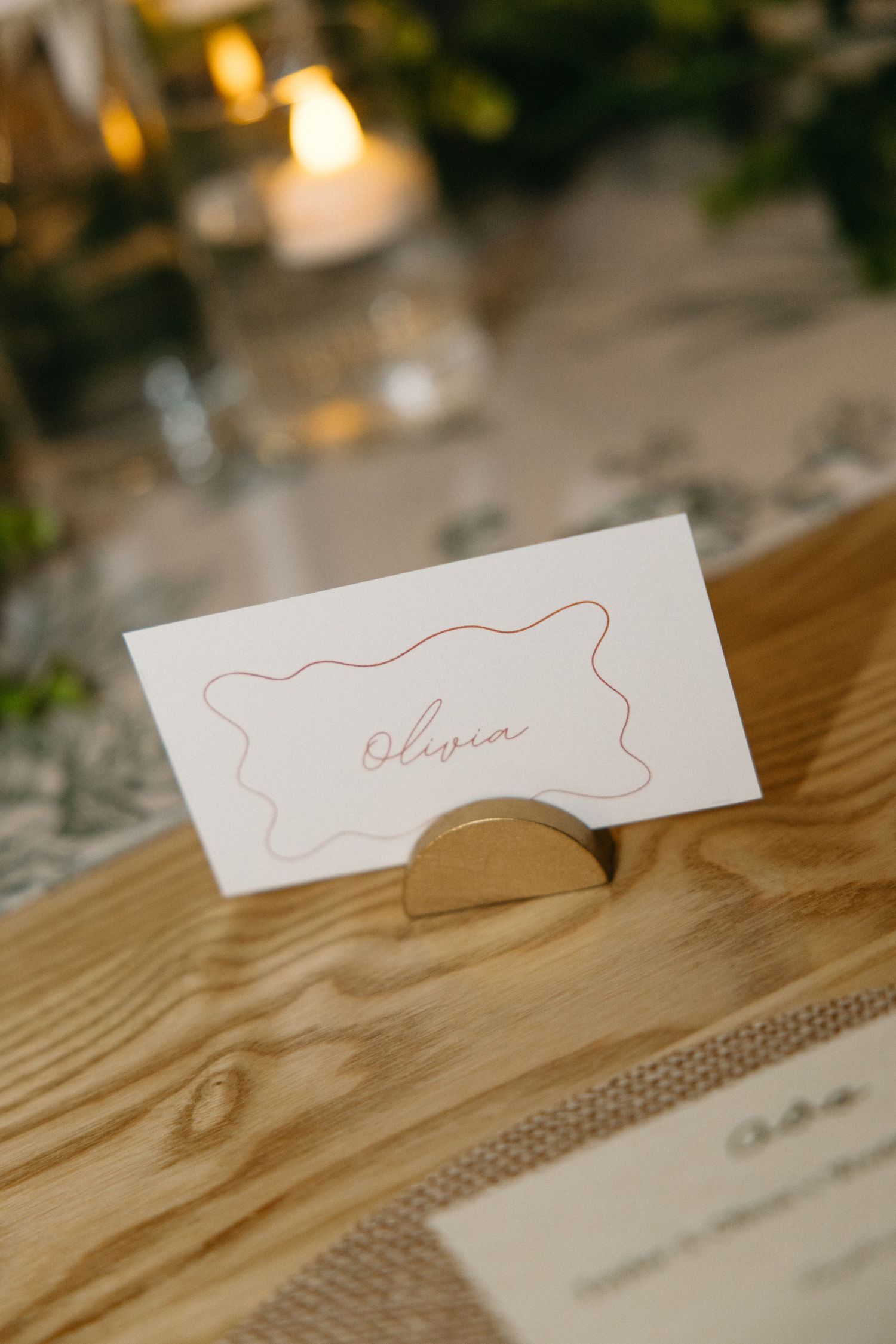 Wedding place card reading Olivia with a wavy border, held in a gold half-moon stand on a rustic wooden table with candles.