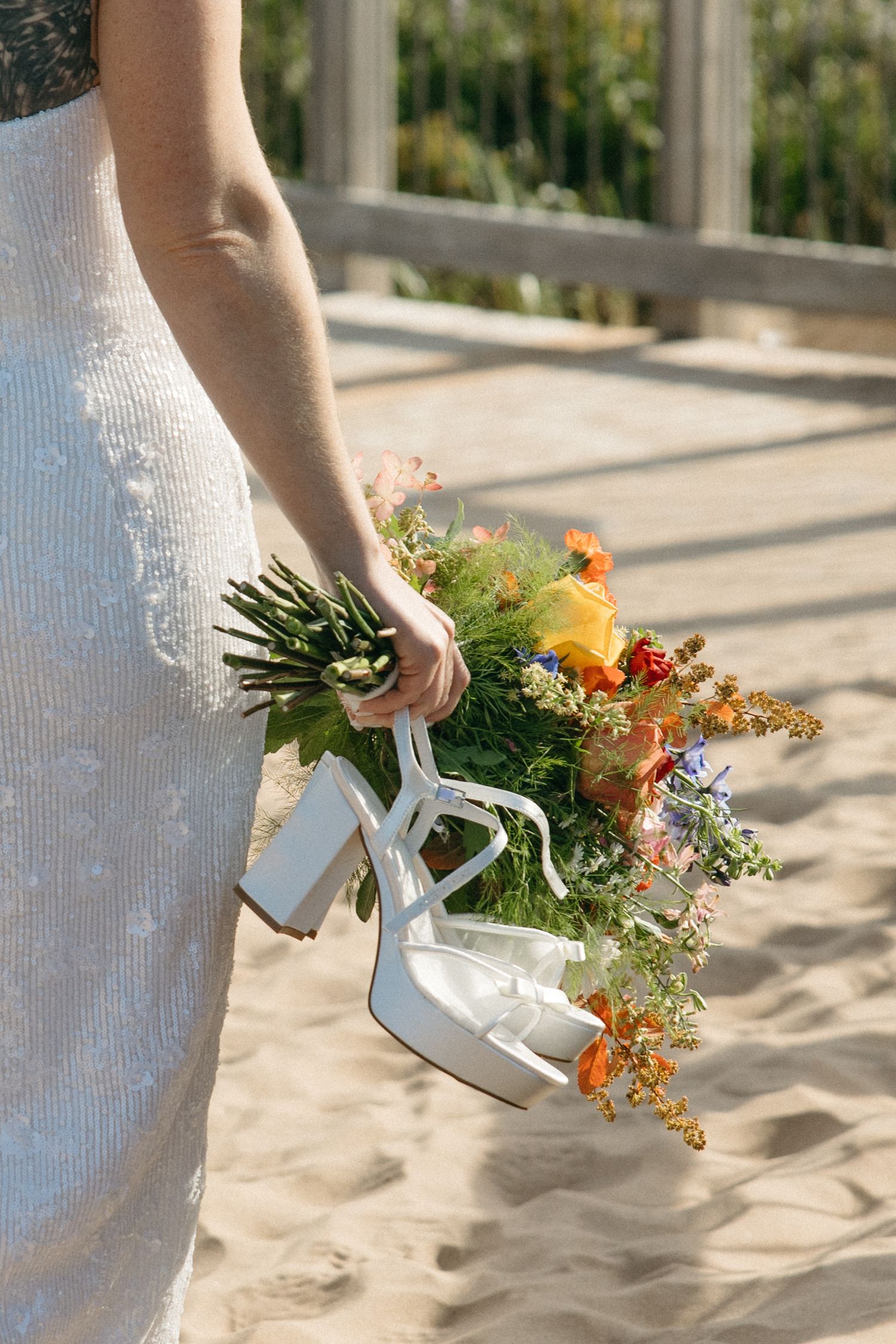 Bride in a white sequined gown holds a colorful wildflower bouquet and white block-heel sandals while standing on a sunny sandy beach.