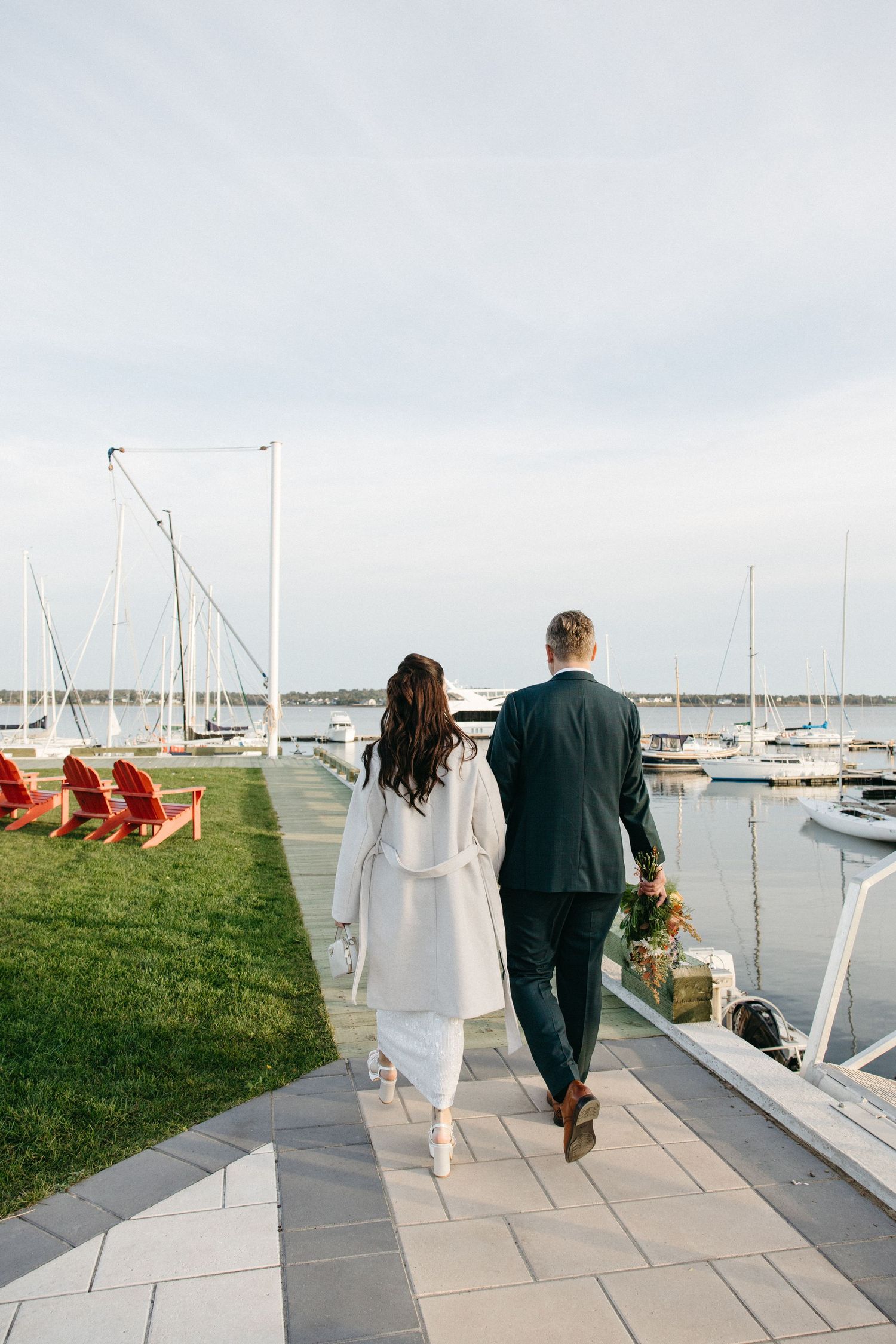 Couple in a white coat and dark suit walk hand in hand along a waterfront marina path past red Adirondack chairs and sailboats.