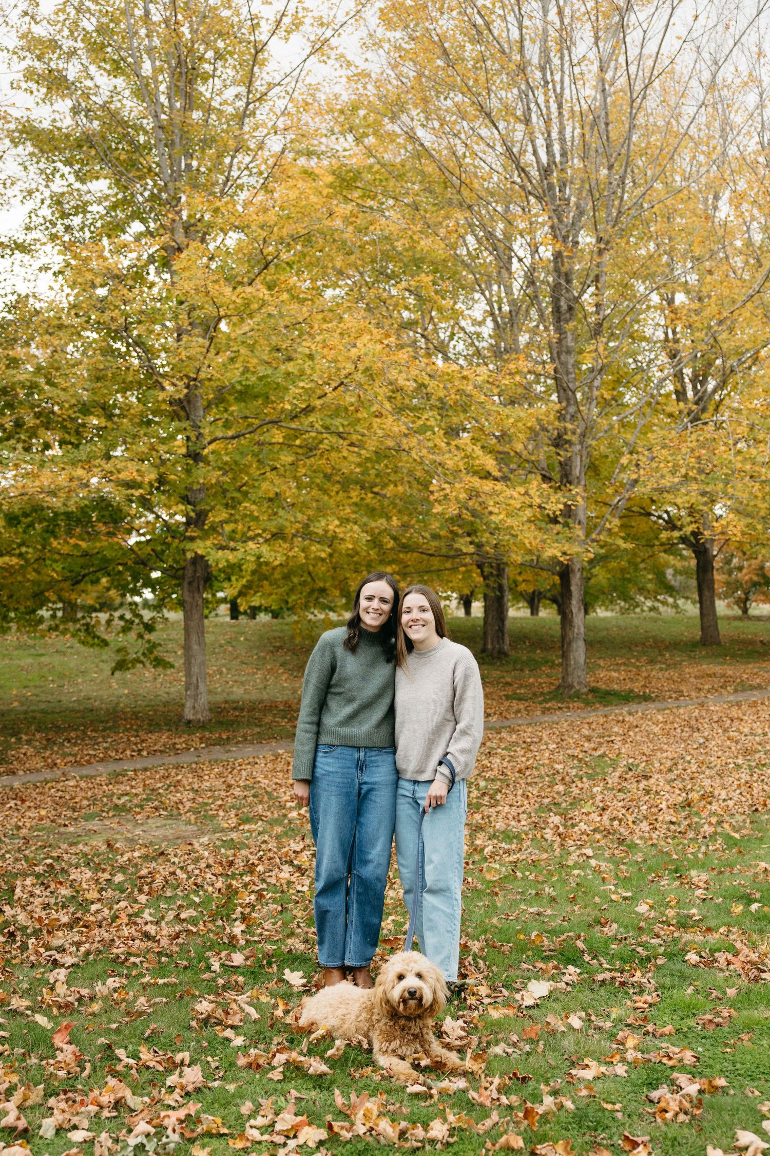 Two women in sweaters and jeans embrace beside their goldendoodle on a leaf-covered lawn beneath golden autumn trees.