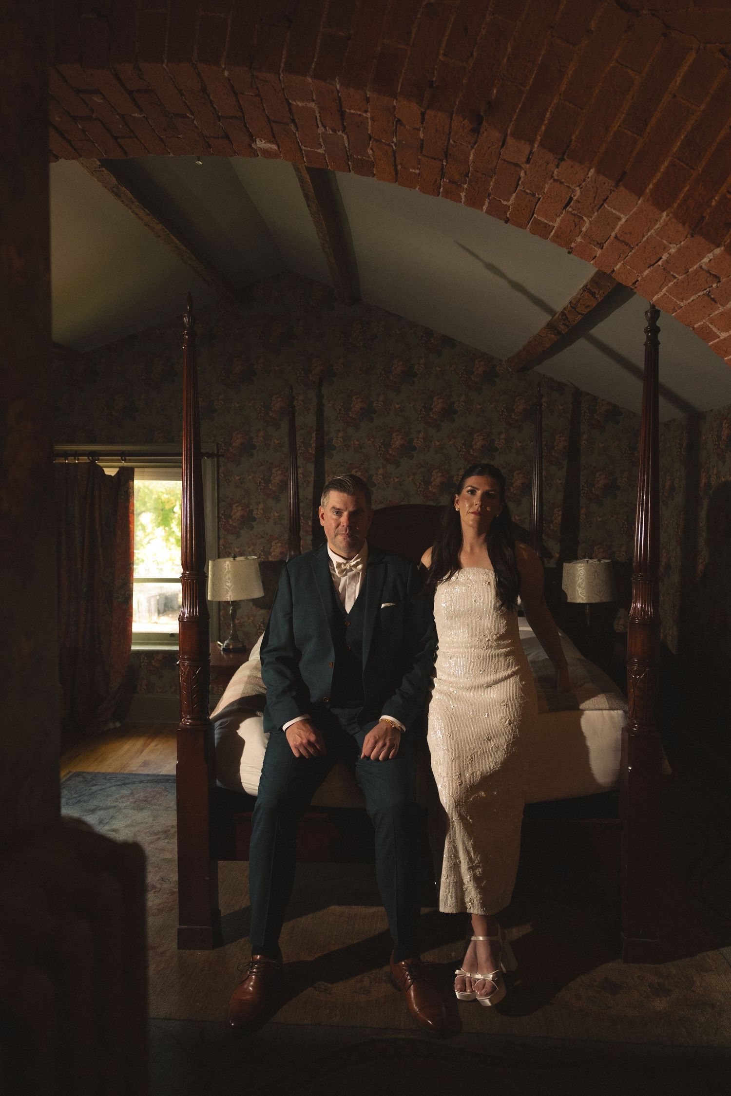 Groom in a navy suit and bride in a lace dress sit on a four-poster bed beneath a rustic brick archway.