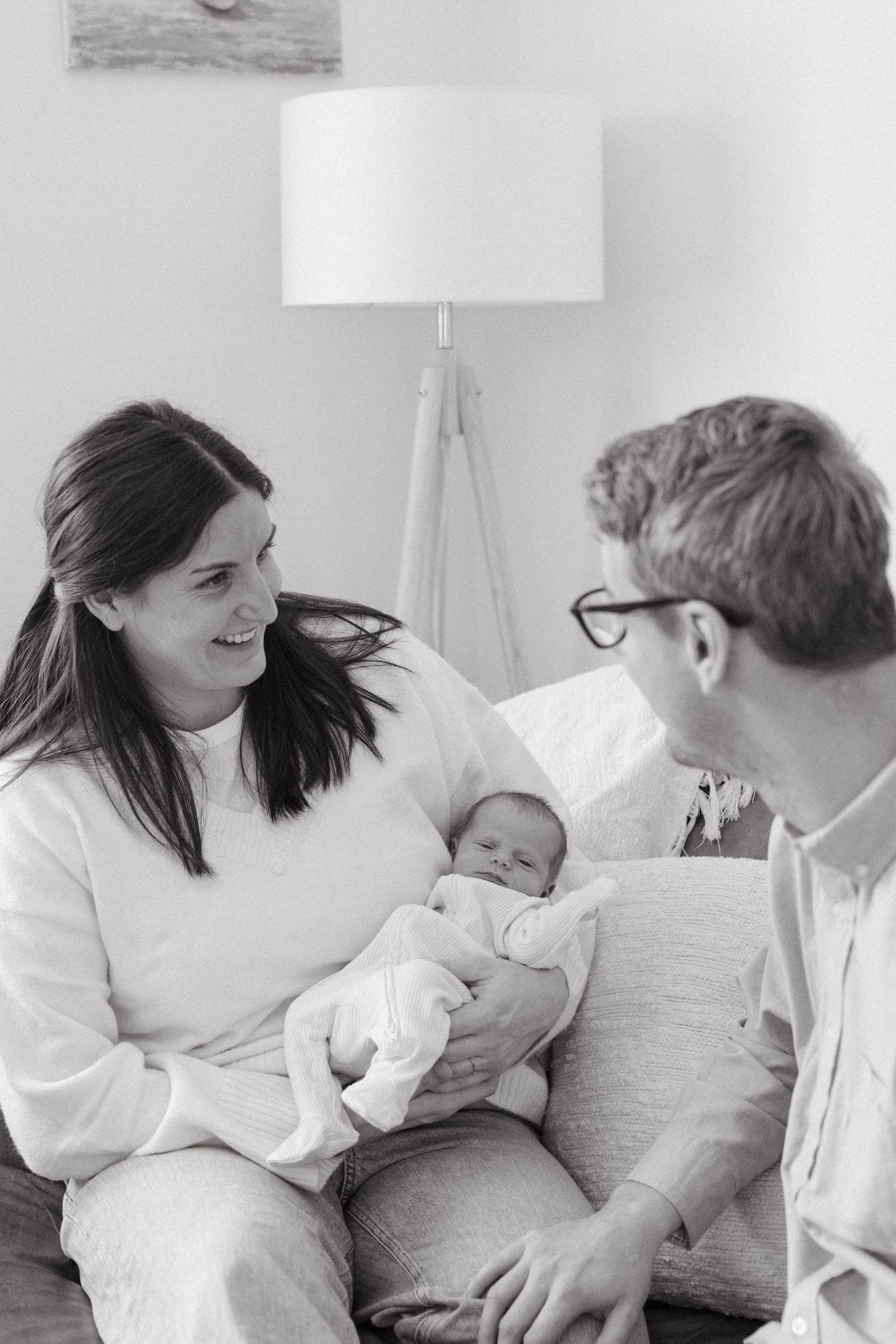 A tender black and white family moment with a newborn baby on a couch near a white floor lamp.