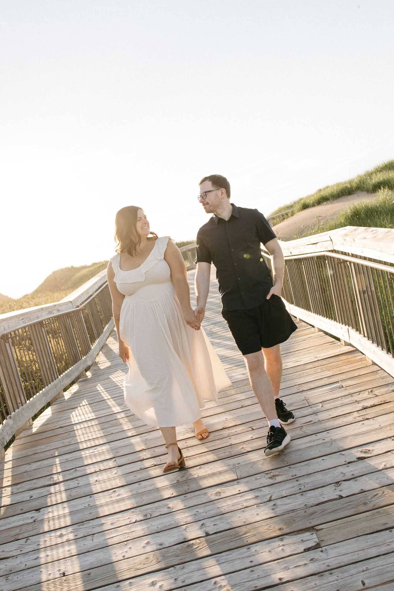 Couple holding hands walking on wooden boardwalk at sunset, woman in flowing white dress and man in black outfit.