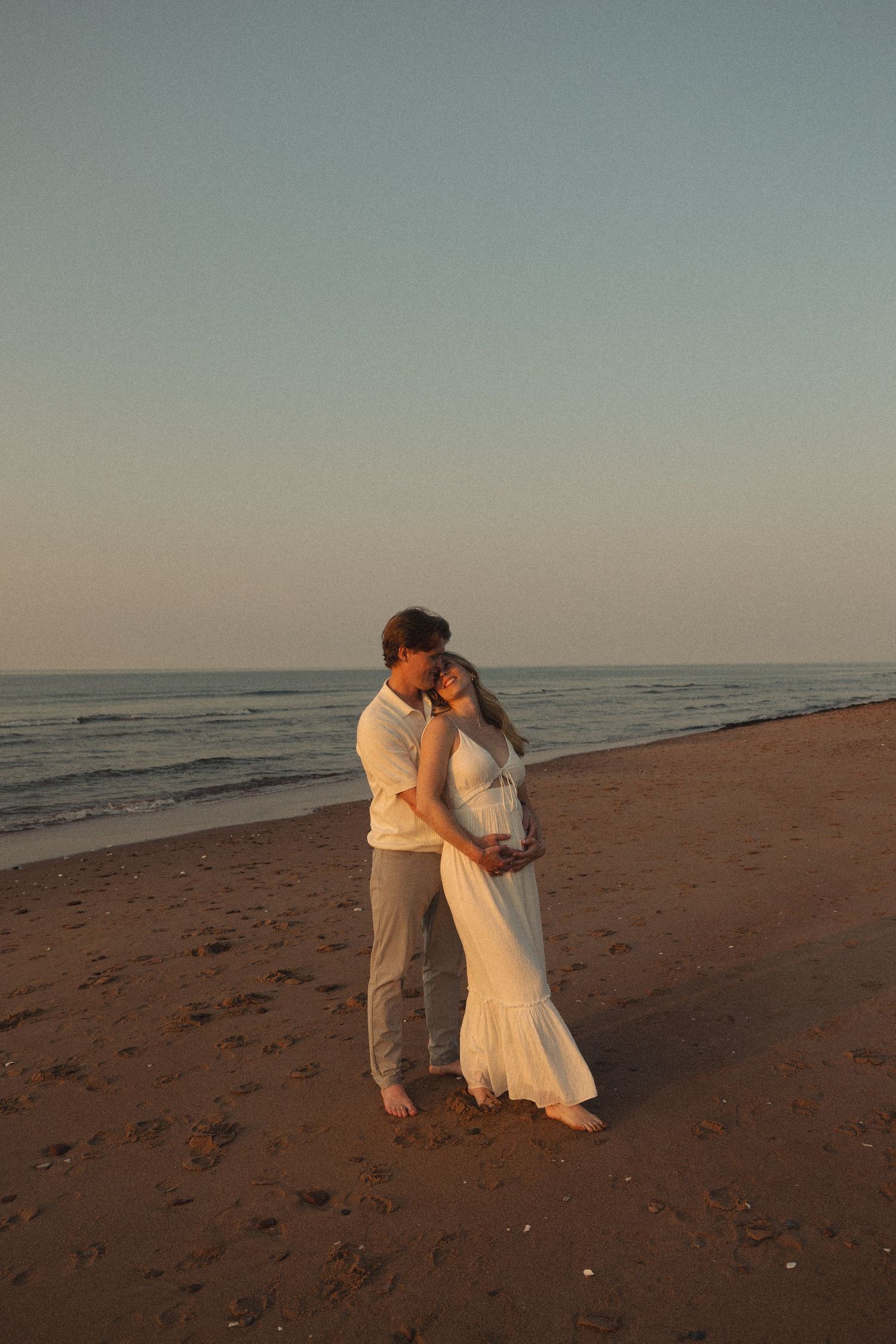 Couple embracing at sunset on a beach, woman in white maxi dress, ocean visible in the background.