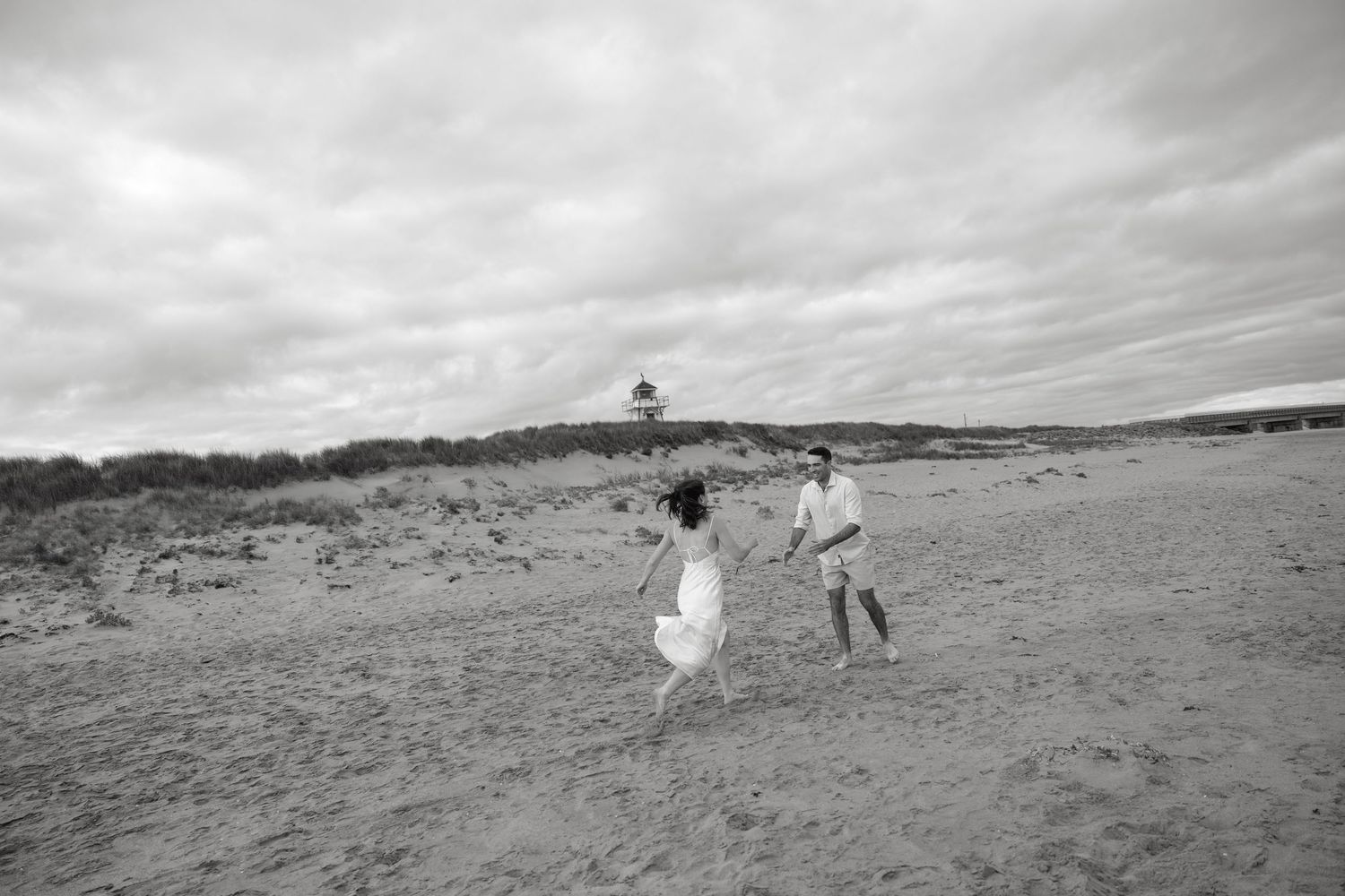 A black and white photo of a couple running together on a beach with a lighthouse in the distance.