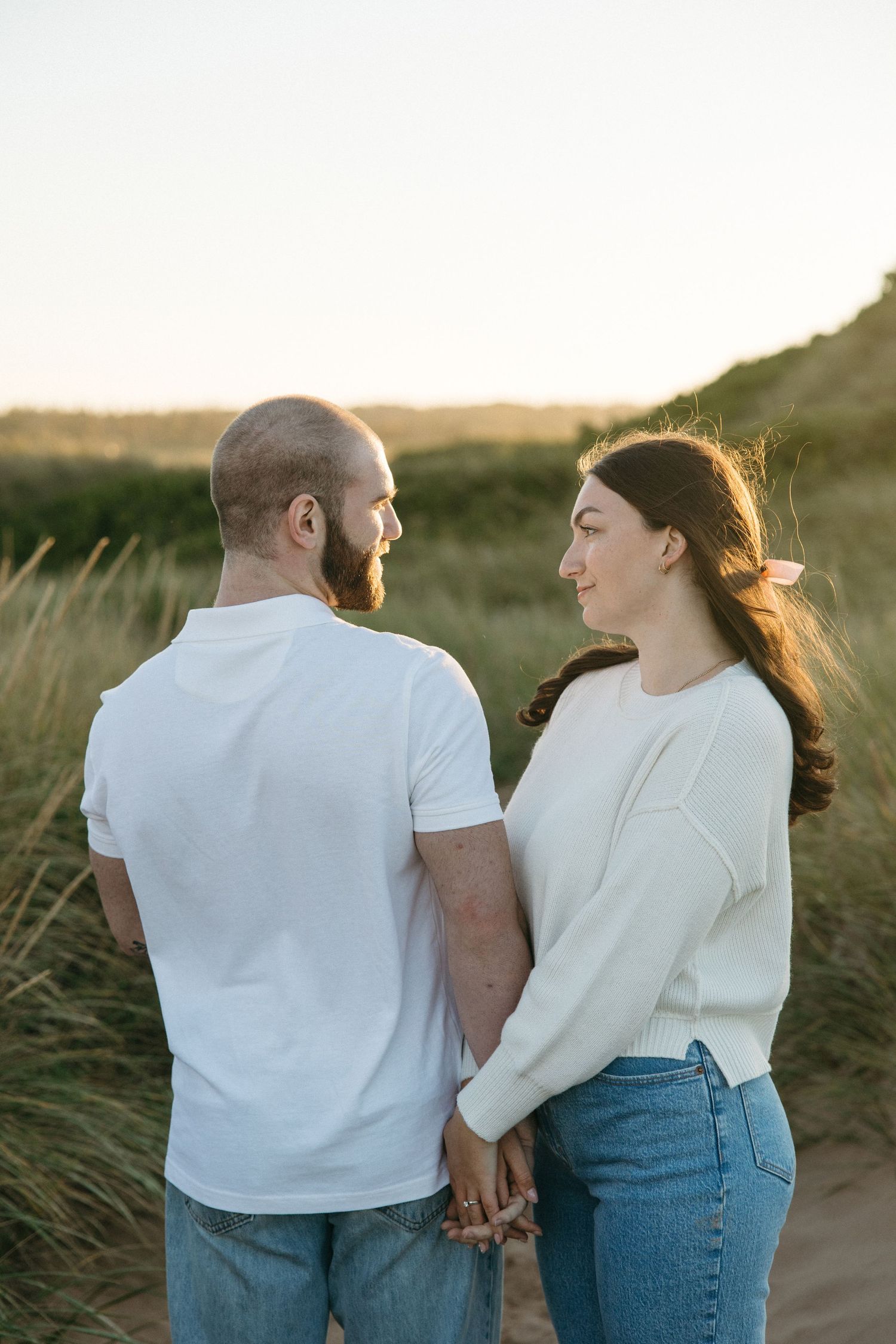 Couple walking hand in hand through beach grass at sunset.