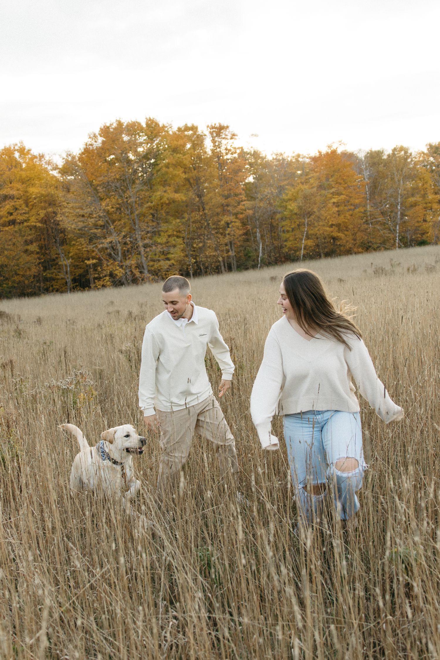 Couple in cream and denim outfits walking with white dog through autumn meadow.