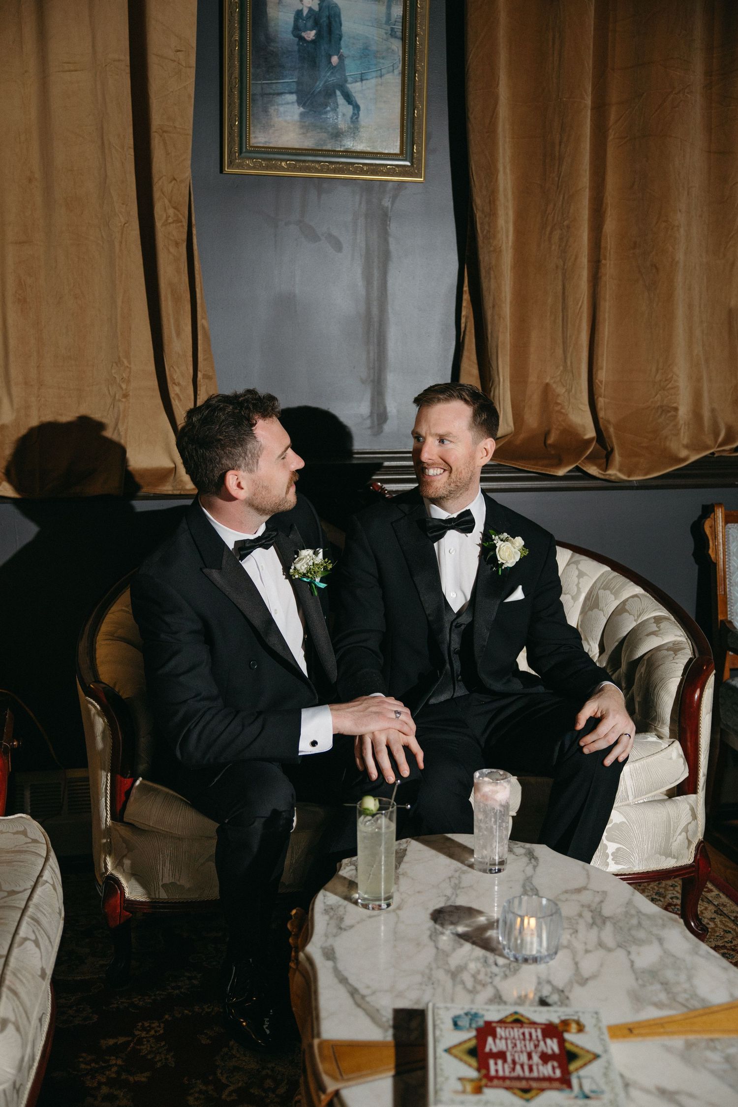 Wedding party members in black suits and bow ties gathered at a dimly lit table for drinks.