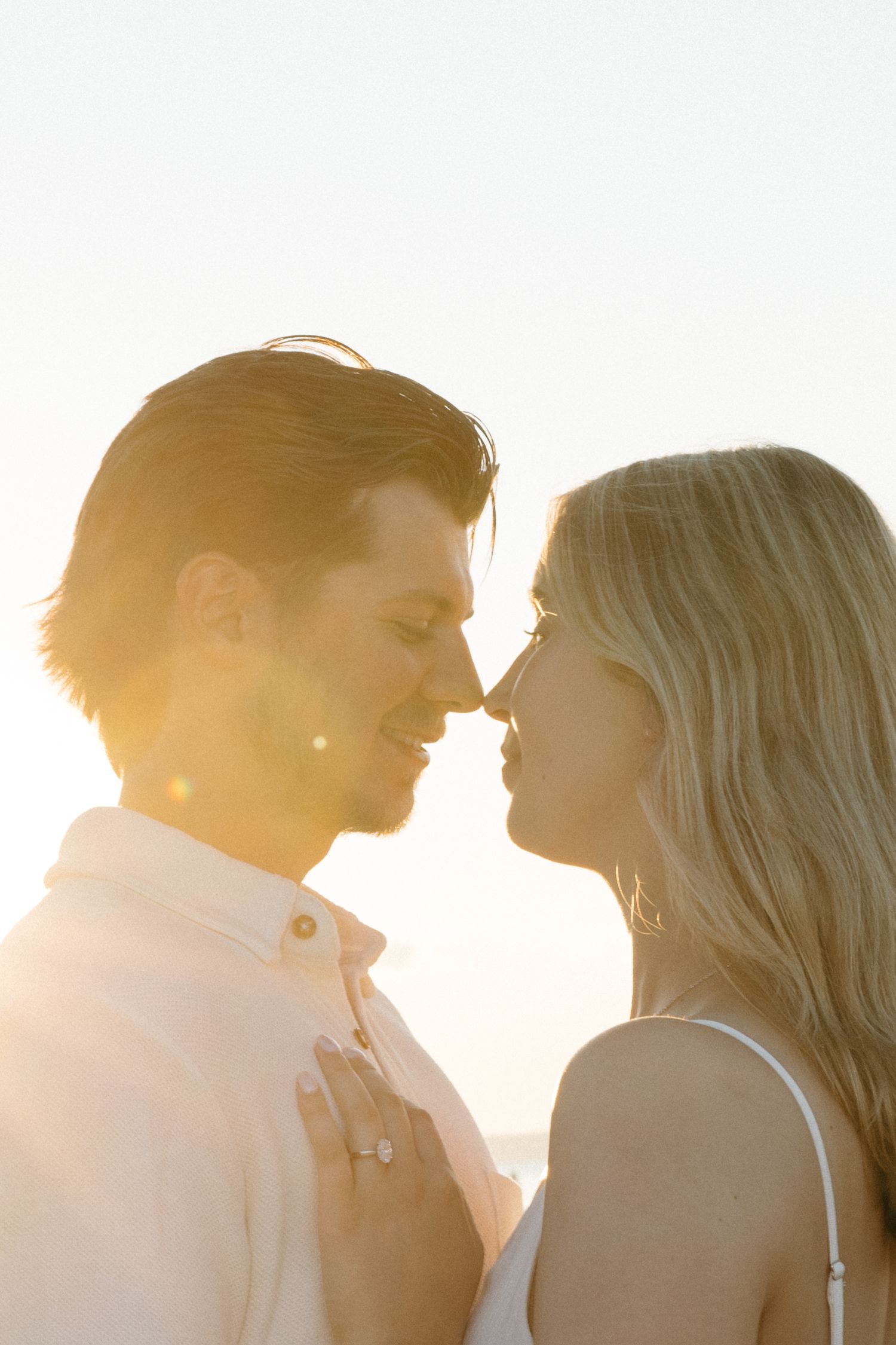 Couple sharing a romantic almost-kiss at golden hour sunset, woman wearing engagement ring, warm lens flare.