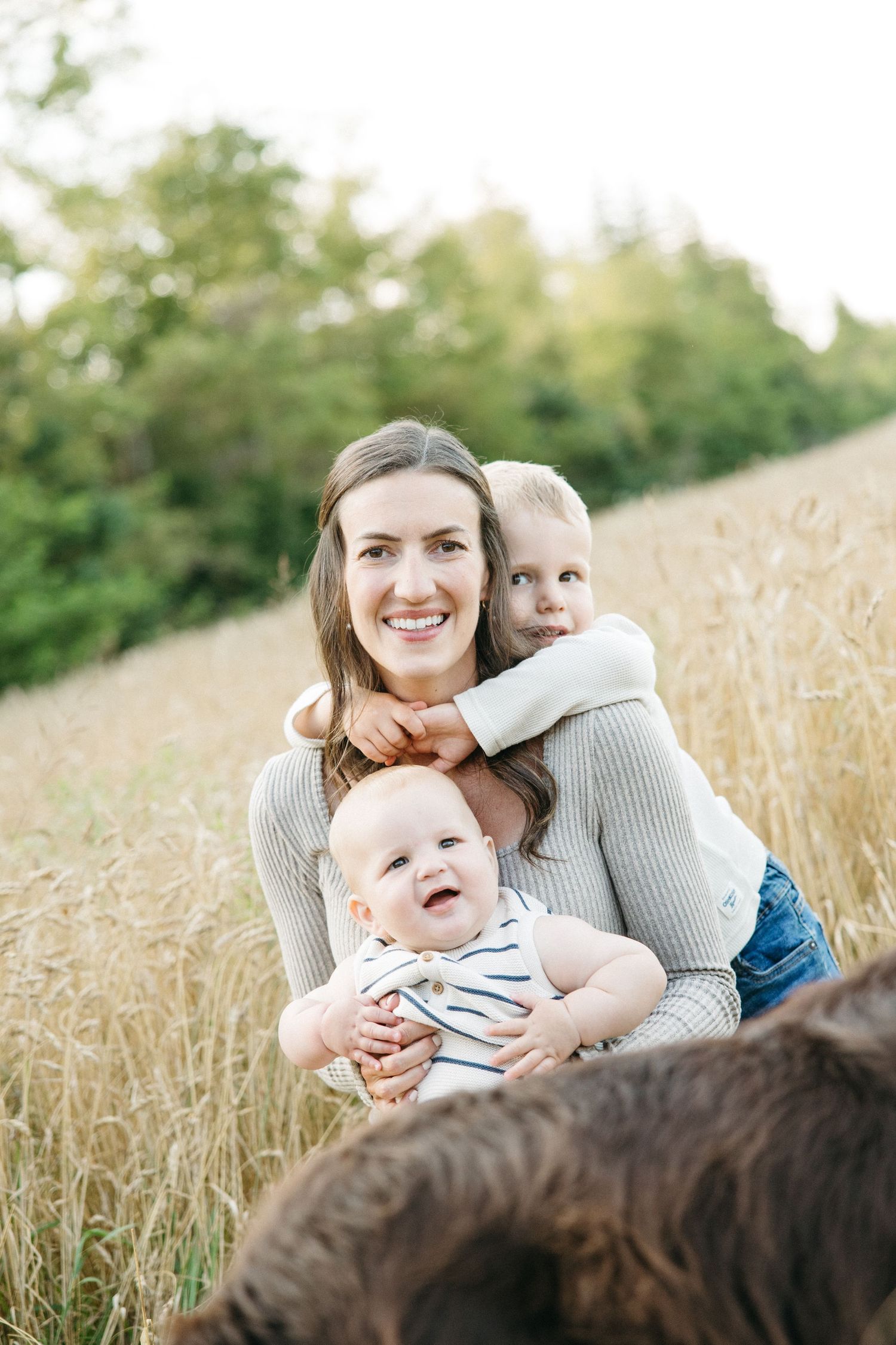 Smiling mother in gray sweater holds baby while toddler hugs her from behind in field.