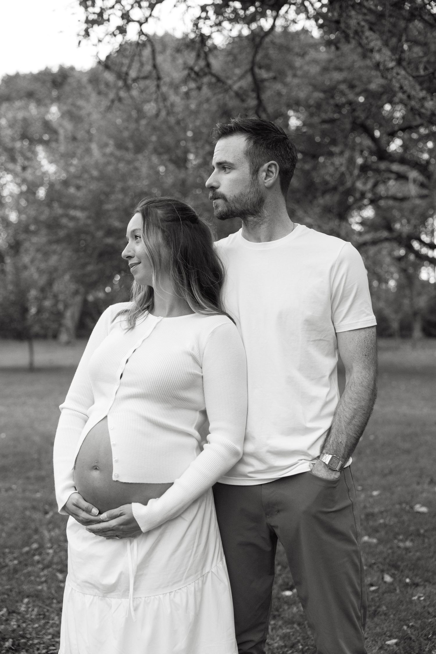 Black and white photo of a pregnant couple standing in a park, the woman cradling her baby bump.