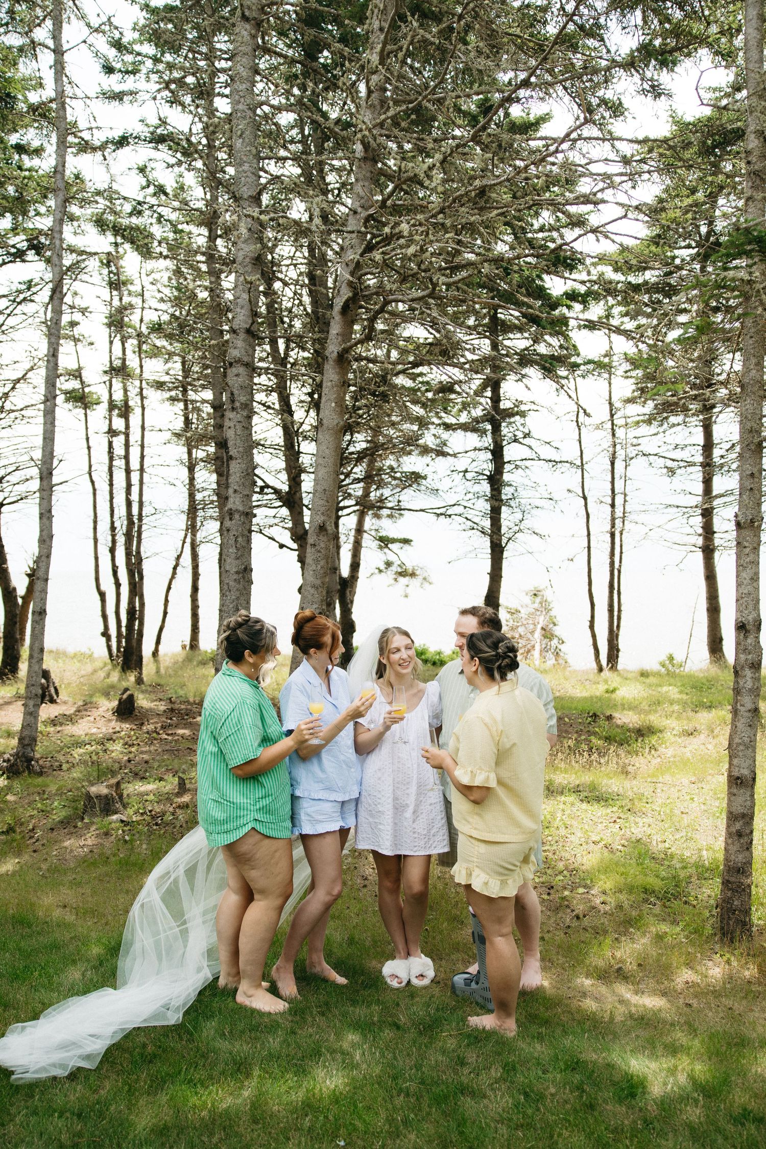 Bridesmaids in pastel pajamas celebrate with bride wearing veil outdoors among tall pine trees.