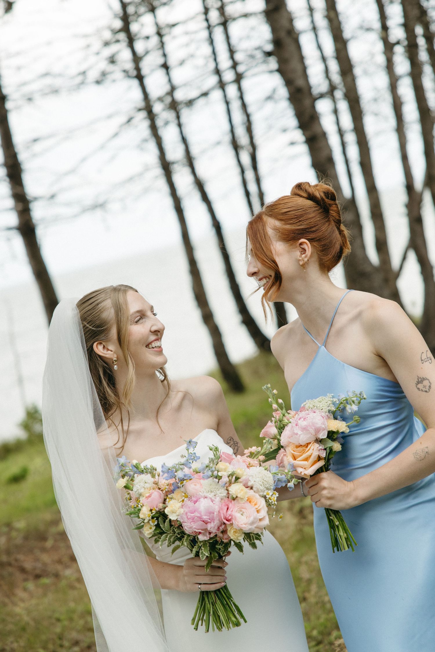 Smiling bride in white gown and veil shares a joyful moment with redheaded bridesmaid in blue satin dress holding bouquets.