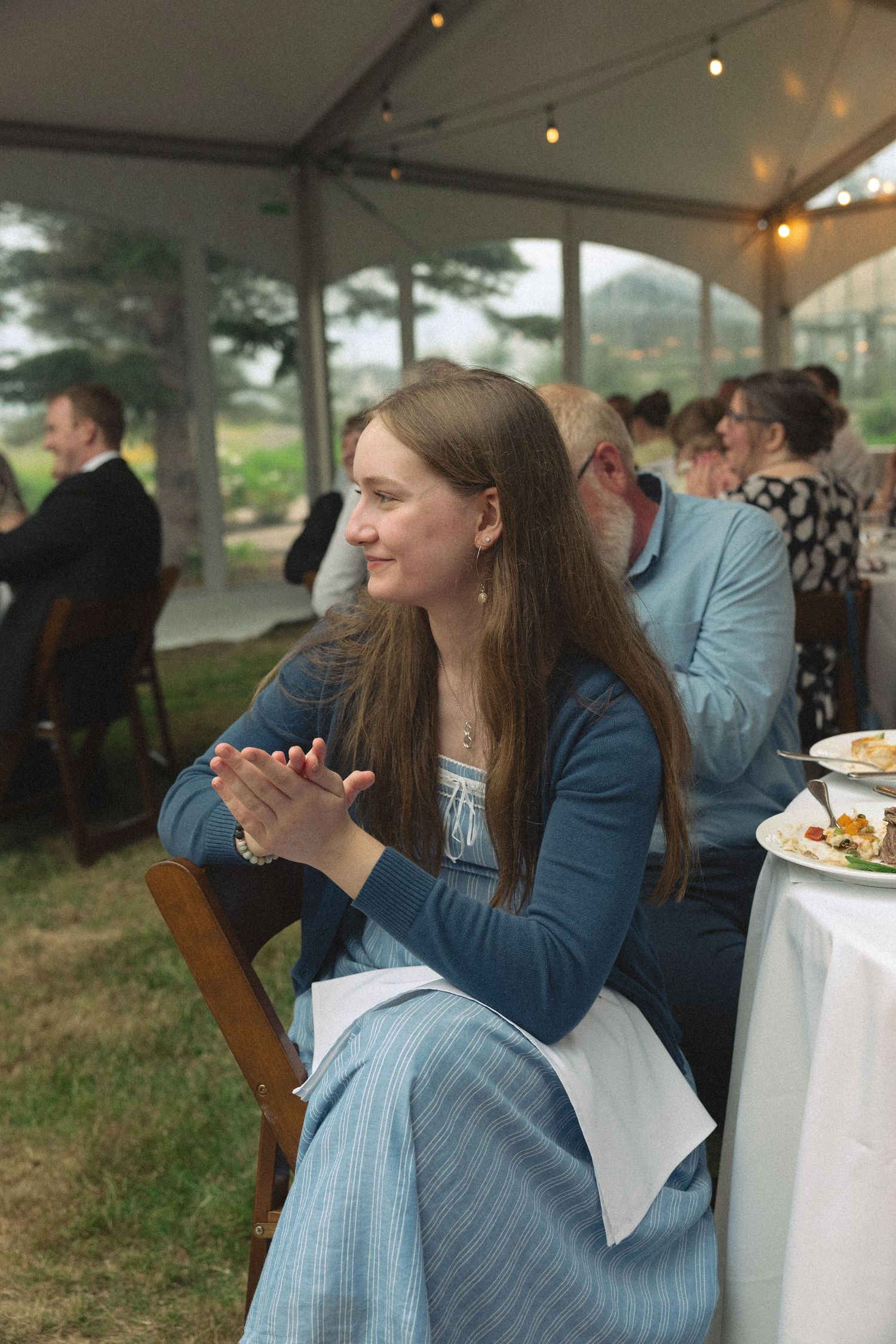 Woman in blue dress clapping at wedding reception under a tent with string lights.