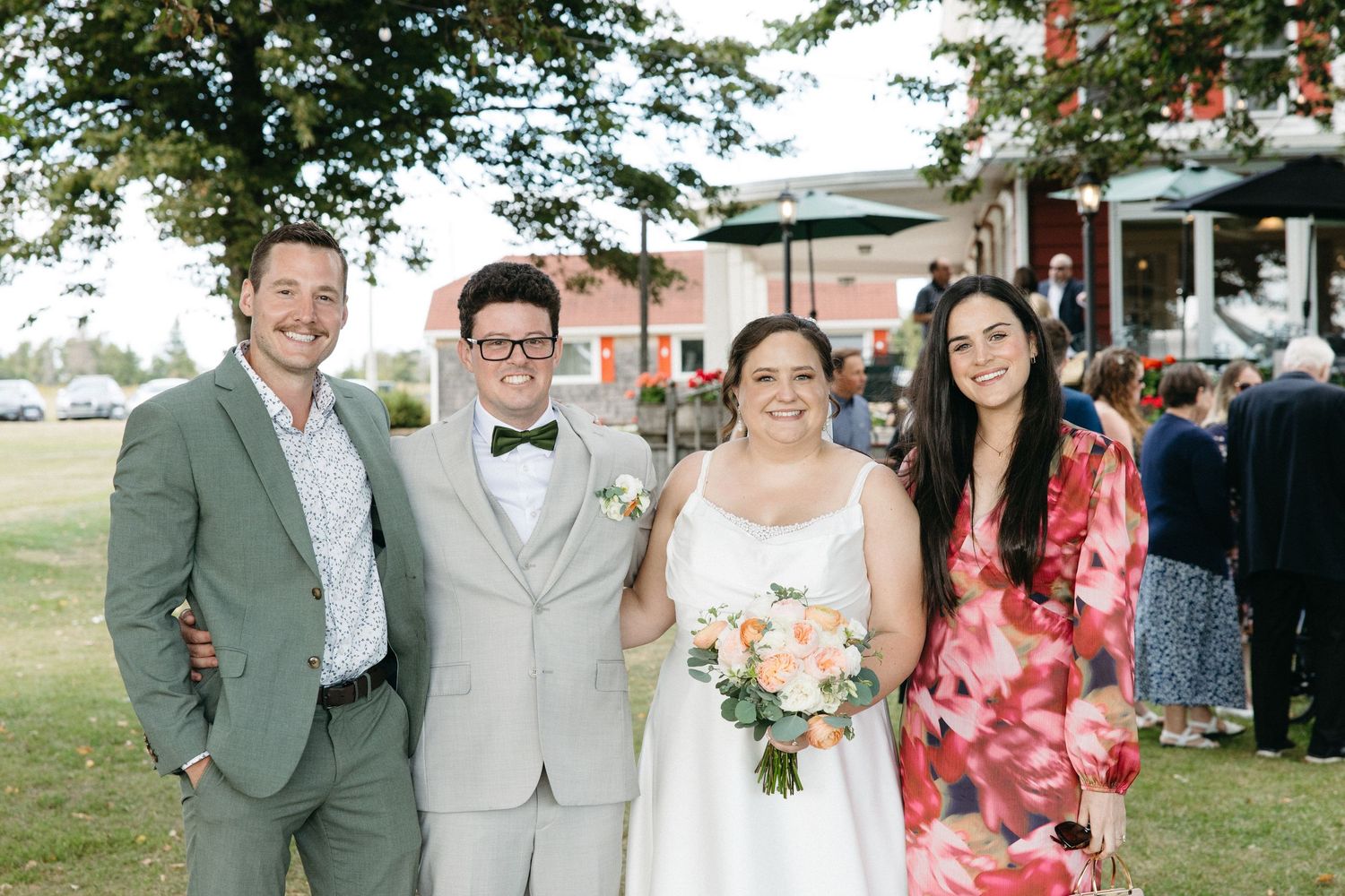 Wedding party of four posing outdoors, bride holding bouquet in white gown with groom in gray suit and bow tie.