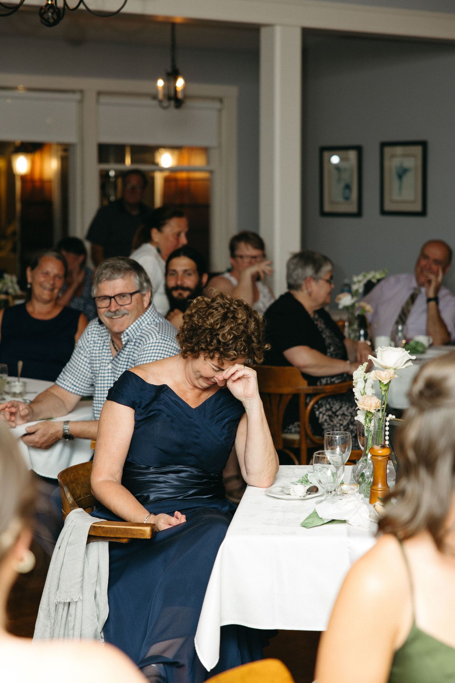 Emotional wedding guest in navy dress wiping tear during reception speeches at elegantly decorated venue.
