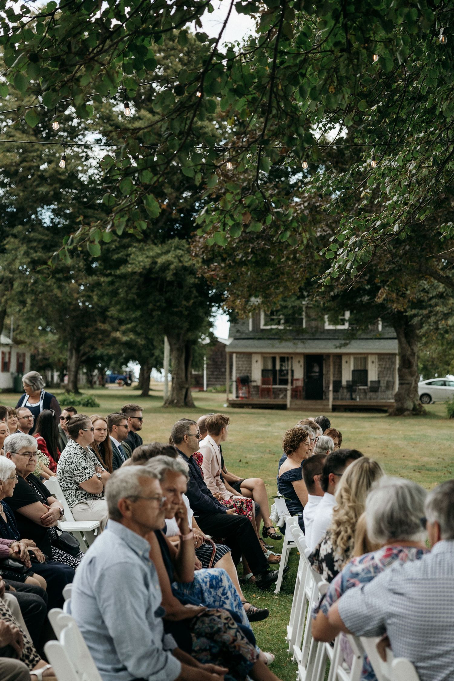 Guests seated outdoors in white chairs for a wedding ceremony under large trees near a historic farmhouse.