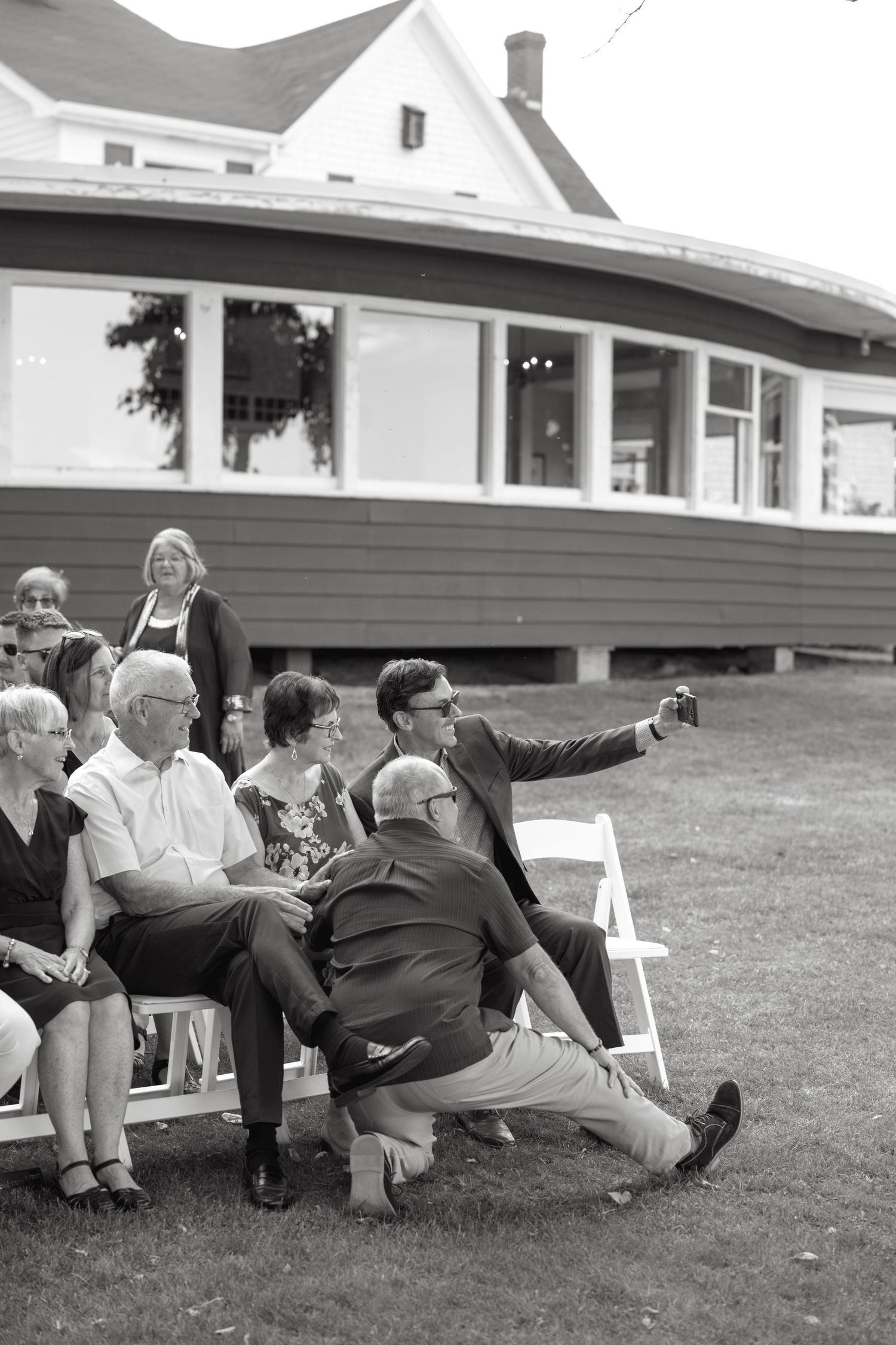Black and white photo of a man taking a selfie with seated wedding guests on a lawn.