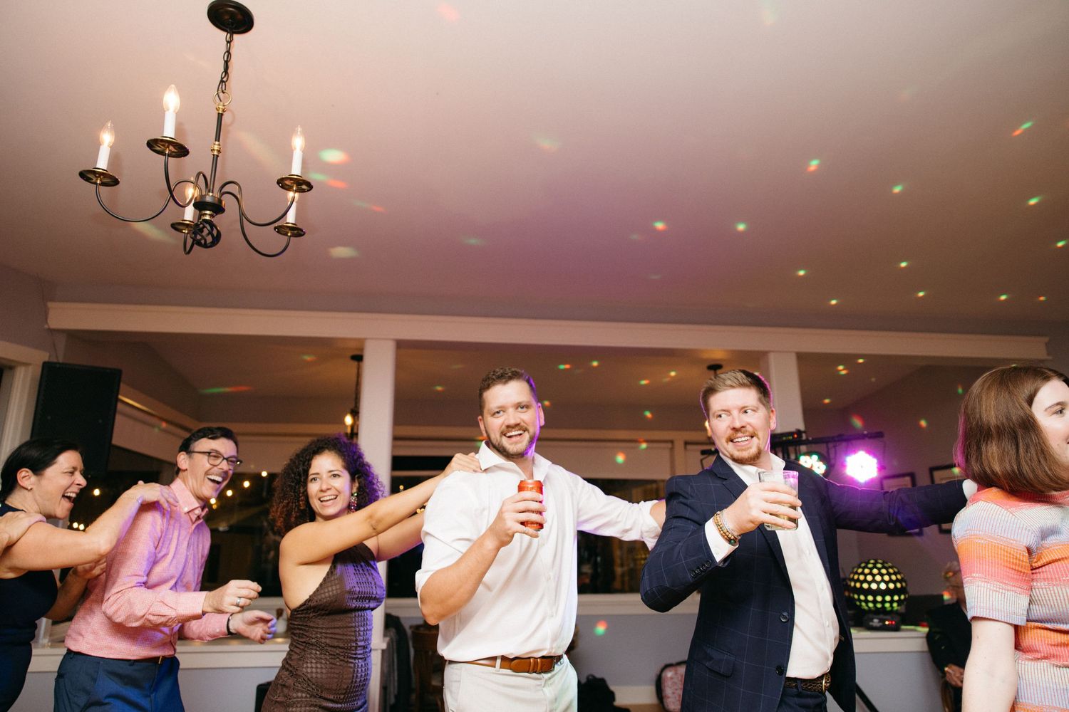Wedding guests dance and celebrate on illuminated reception dance floor holding drinks.