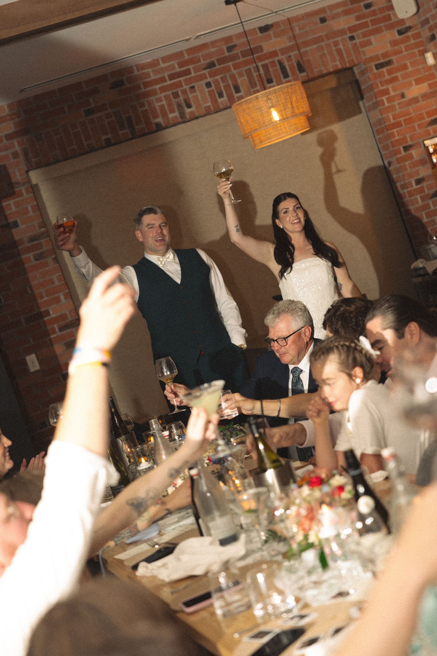 Bride and groom raise their glasses in a toast at a wedding reception while seated guests look on at a long table in a brick-walled venue.