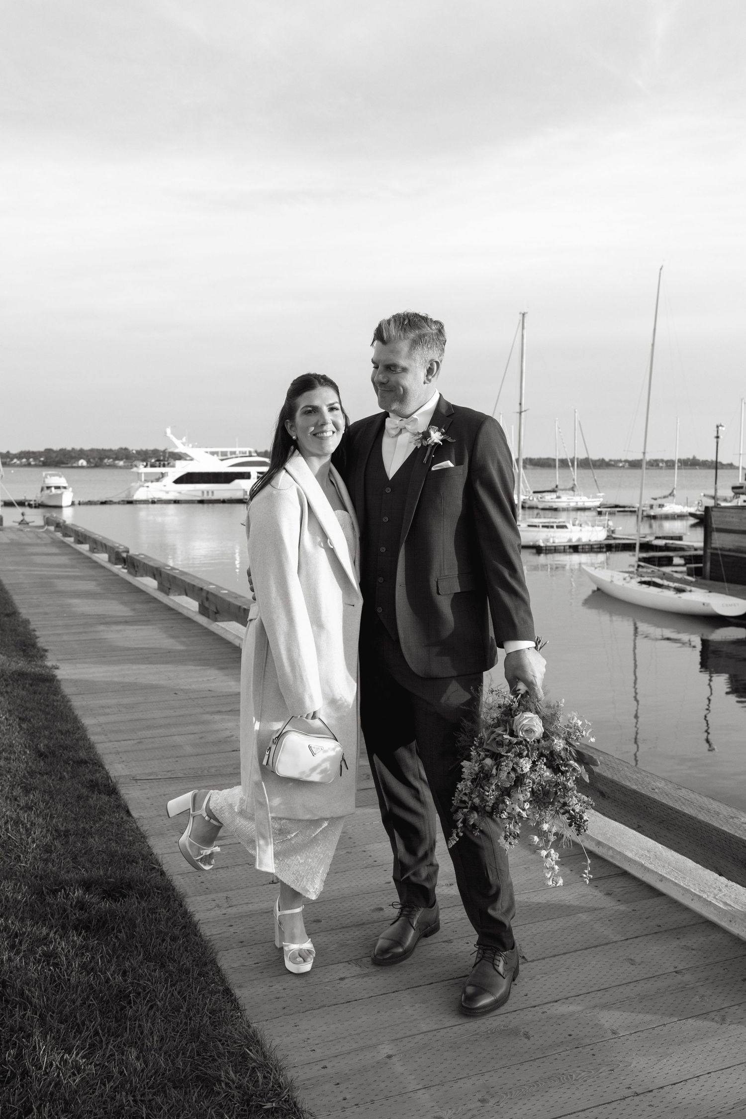 Black and white photo of a smiling bride in a white coat and groom in a vest holding a bouquet on a waterfront dock with sailboats.