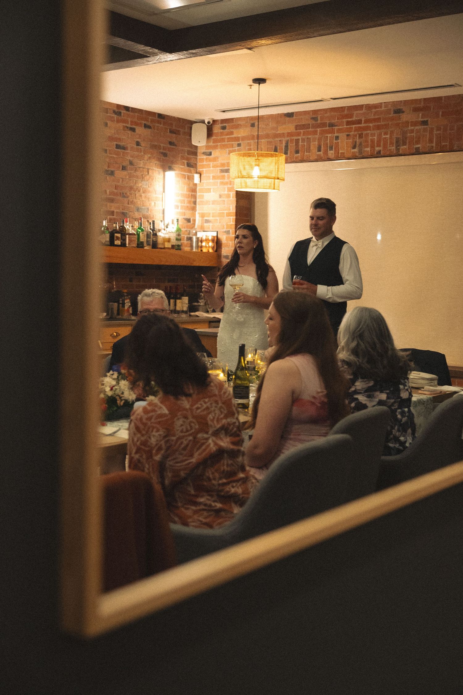A woman and a man stand holding drinks to give a toast at a candlelit restaurant gathering with guests seated at tables nearby.