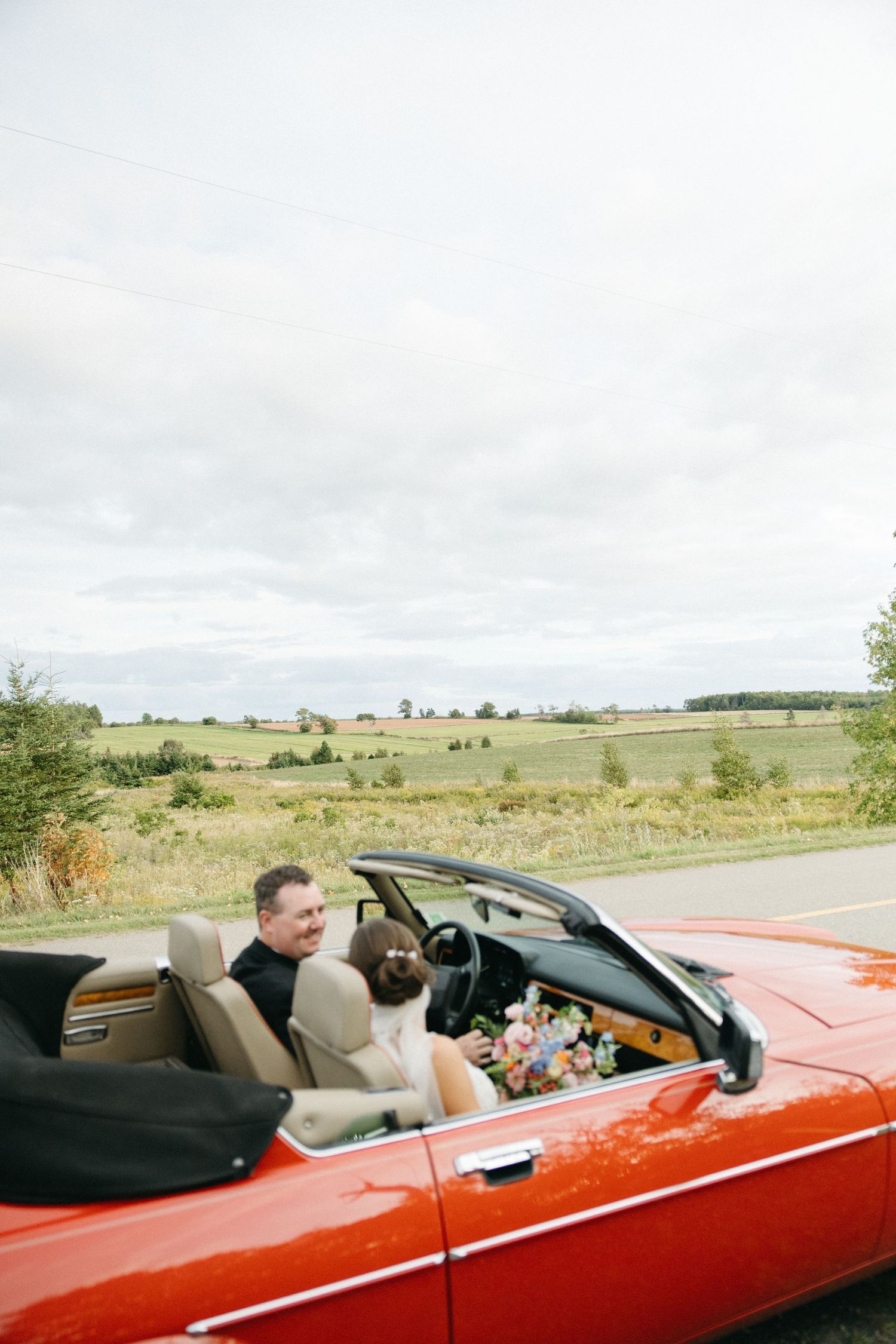 Newlyweds riding in a red convertible car through countryside landscape.