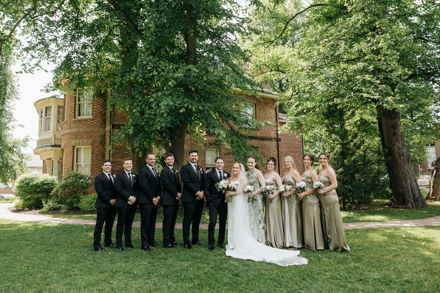 Wedding party poses on the lawn in front of a historic brick mansion on a sunny day.