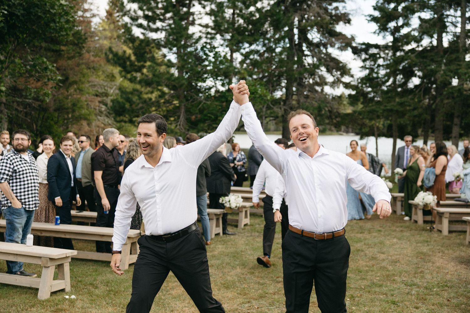 Wedding guests celebrating outdoors with raised arms under tall pine trees.