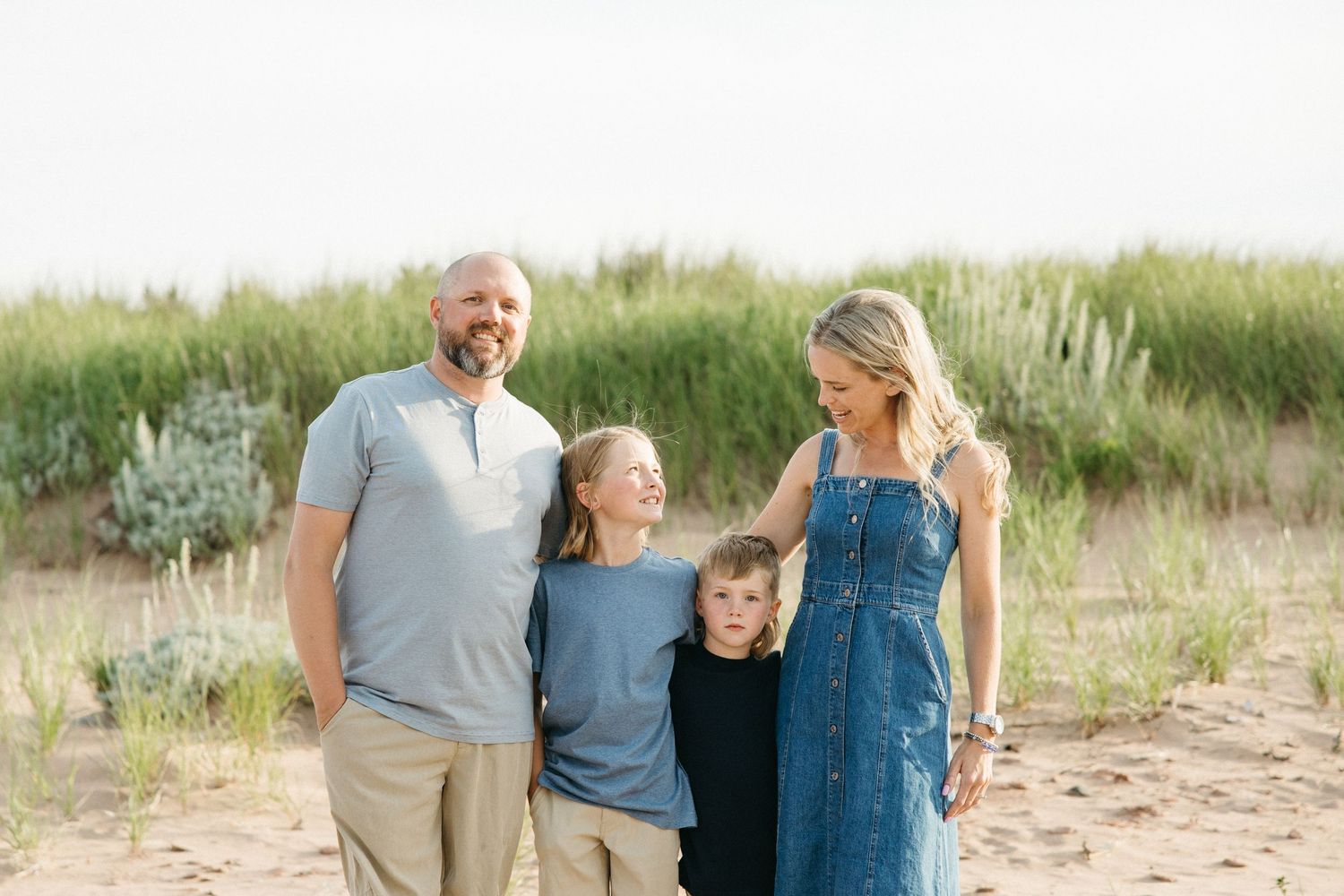 A smiling family of four poses together on a sandy beach with green dune grass behind them.