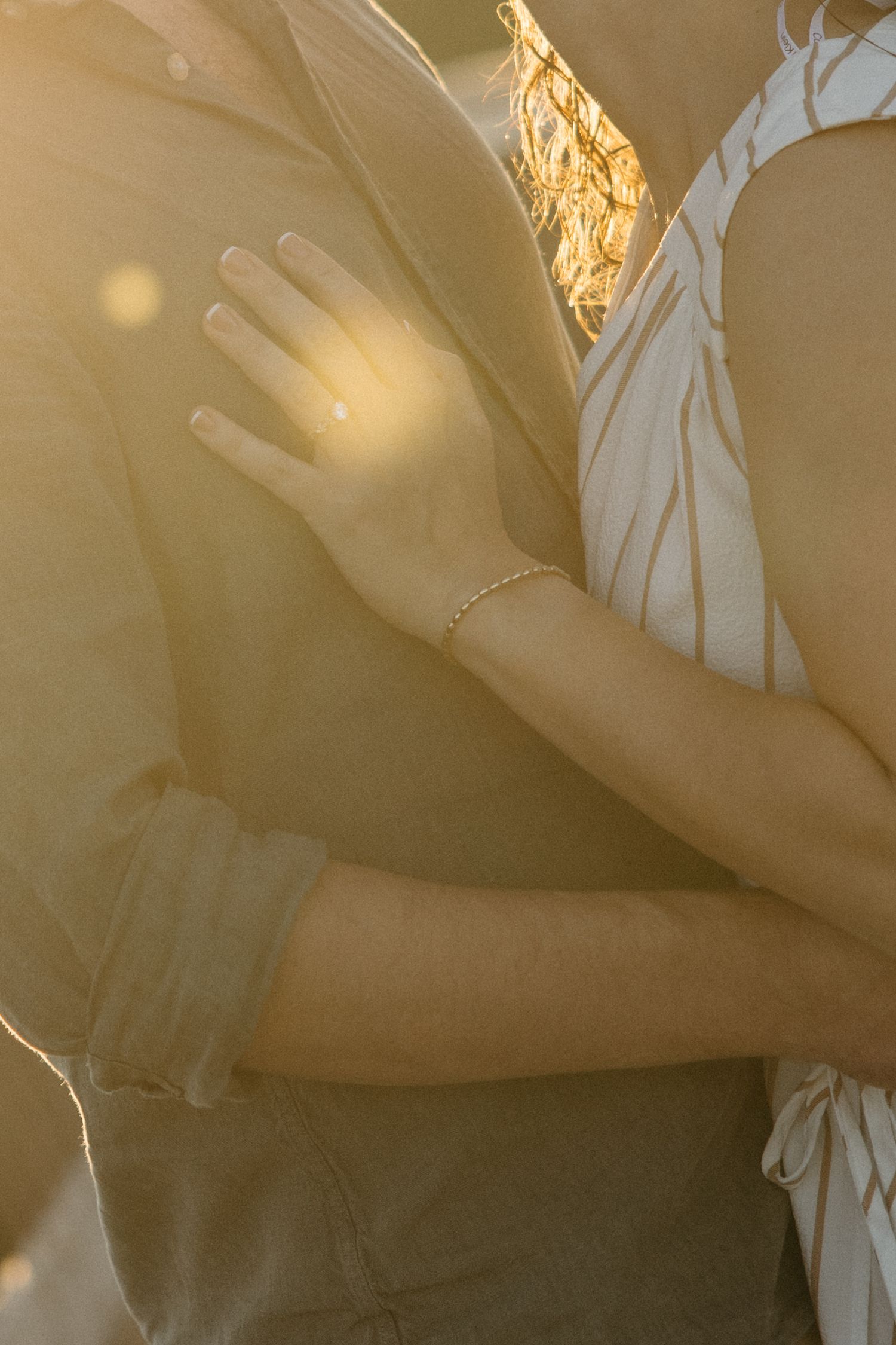 Two people embracing in golden sunset light, woman wearing a delicate gold bracelet and engagement ring.